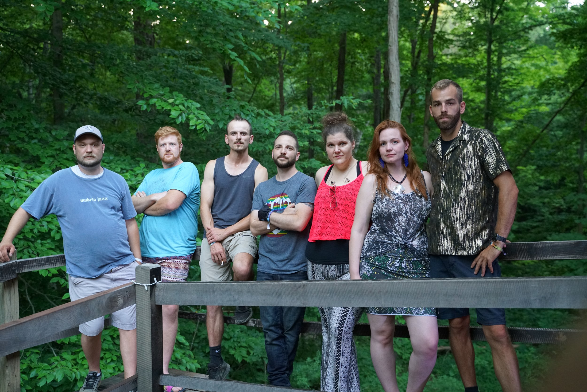 A group of seven people standing on a wooden deck in a forest setting. They are casually dressed, with green trees in the background.