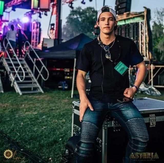 Young man wearing a black T-shirt, ripped jeans, and sunglasses, standing outdoors near sound equipment at a music event or festival.
