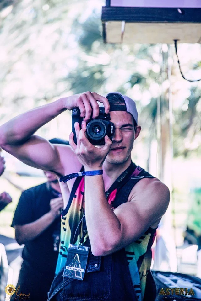 Young man in colorful sleeveless shirt taking a selfie with a camera, wearing a backwards cap and a wristband, outdoors with blurred trees in the background.