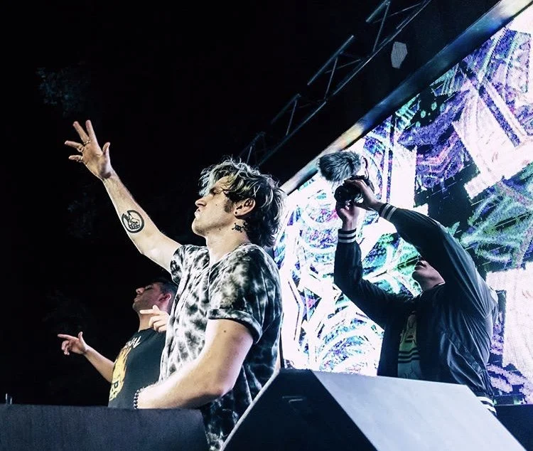 Three young men DJing at a music event with a large LED screen displaying colorful visuals in the background.