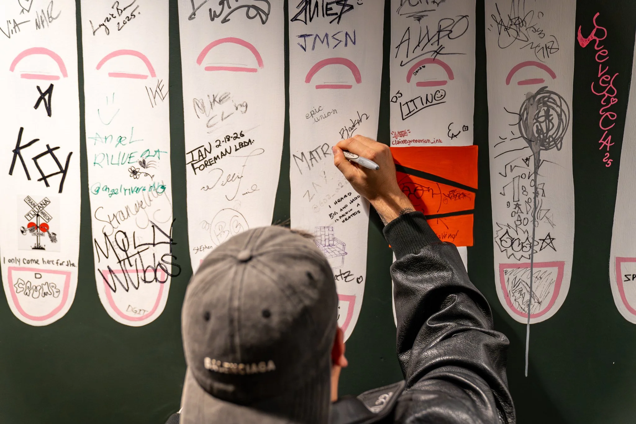 People signing or writing on large sheets of paper that are attached to a green wall, with signatures, drawings, and handwritten notes visible.