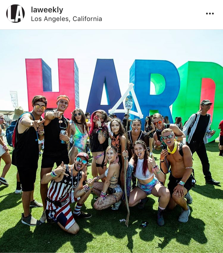 Group of young people at an outdoor event in front of colorful LA letters, posing happily, some pointing at the camera, wearing summer clothing and accessories.