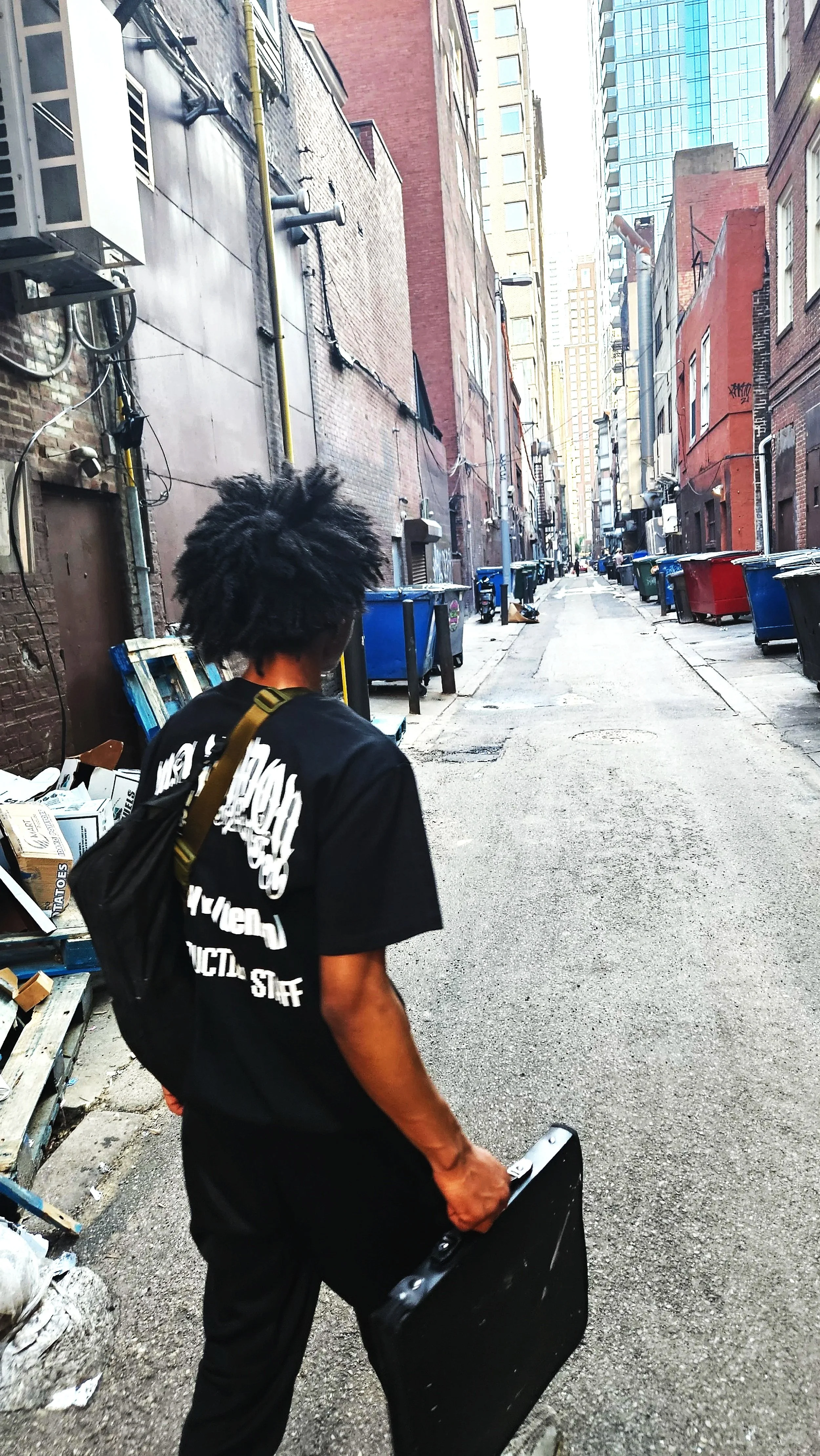 Person with dreadlocks carrying a black case walking through a narrow urban alleyway lined with trash bins.