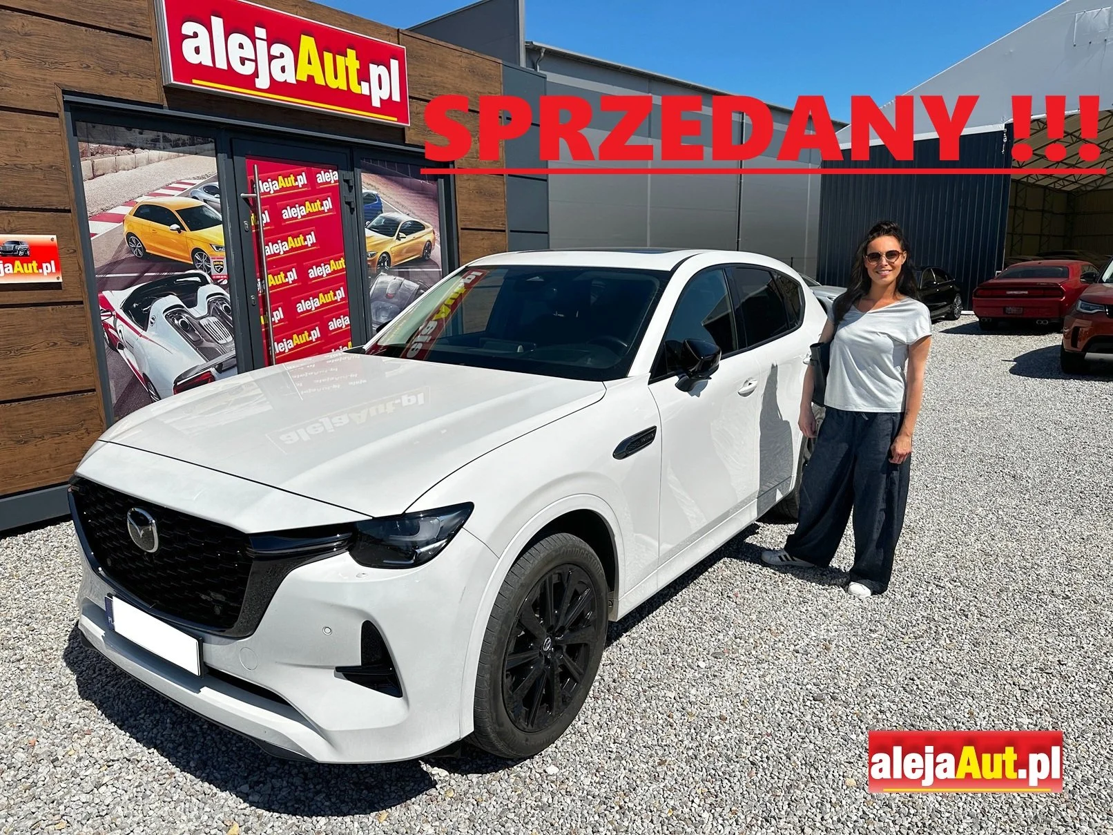 A woman standing next to a white Mazda car outside a dealership with a sign that reads 'SPRZEDANY!!!' in red letters. The dealership has a sign that says 'alejaAut.pl' and display windows showcasing several cars.