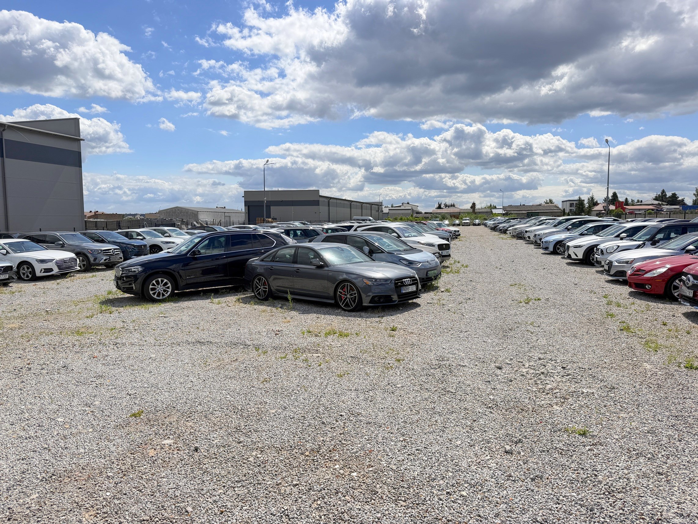 A large outdoor parking lot with rows of parked cars, mostly sedans and SUVs, on a gravel surface. In the background, there are industrial buildings and a partly cloudy sky.
