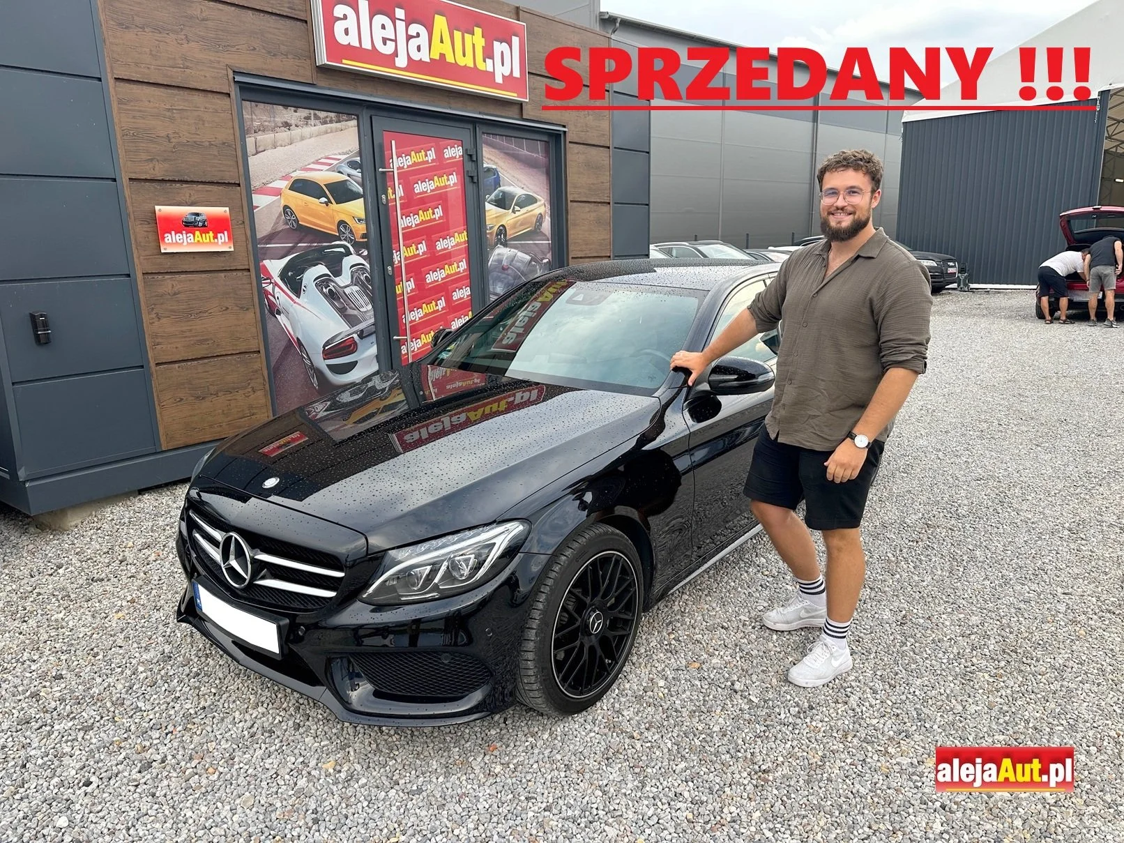 A man smiling and standing next to a black Mercedes-Benz car outside a dealership with a sign that says 'SPRZEDANY' indicating the car is sold. The dealership has posters with various colorful cars and the website alejaAuto.pl. The man is wearing a gray shirt, black shorts, white sneakers, and striped socks.