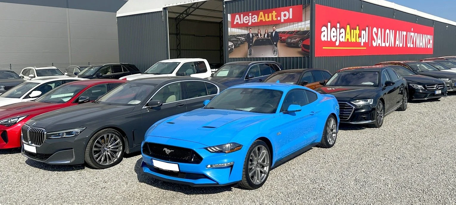 A lot of cars parked outside an auto dealership with a red billboard that reads 'Aleja Aut.pl SALON AUT UŻYWANYCH' and an advertisement showing a group of people with cars.