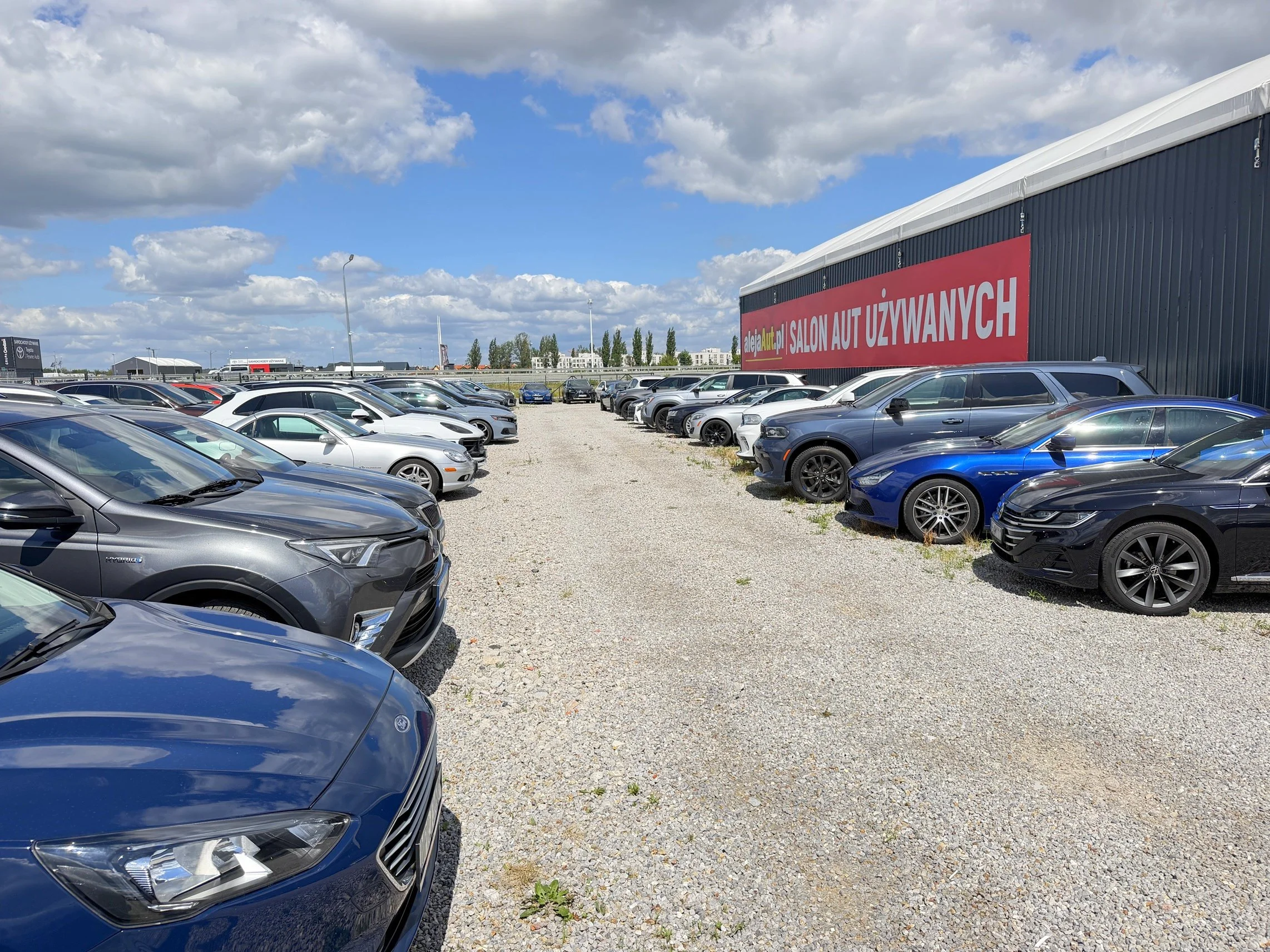 Cars parked in a gravel lot next to a building with a large red sign that reads "Salon Aut Używanych." The sky is mostly cloudy with patches of blue, and there are trees and additional buildings in the distance.