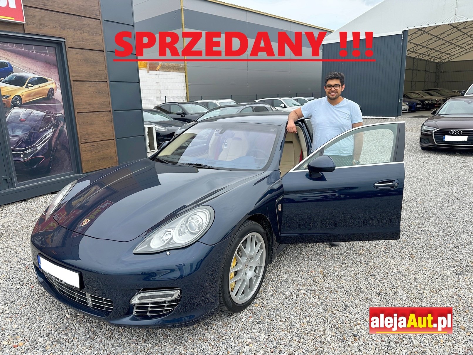 A man standing next to a dark blue Porsche sports car with the door open at a car dealership lot. The word 'SPZEDANY' in red with exclamation marks is overlaid at the top, and a red and yellow logo 'alejaAuto.pl' is at the bottom right corner.