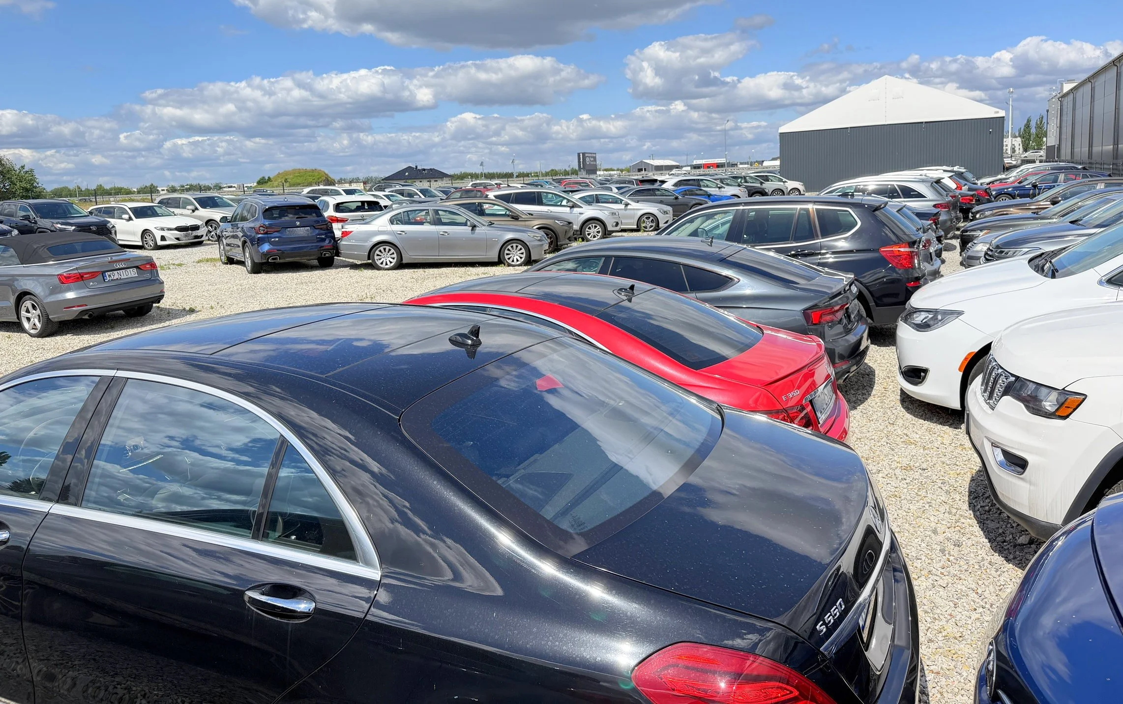 A large outdoor parking lot filled with numerous parked cars of various makes and models, under partly cloudy skies.