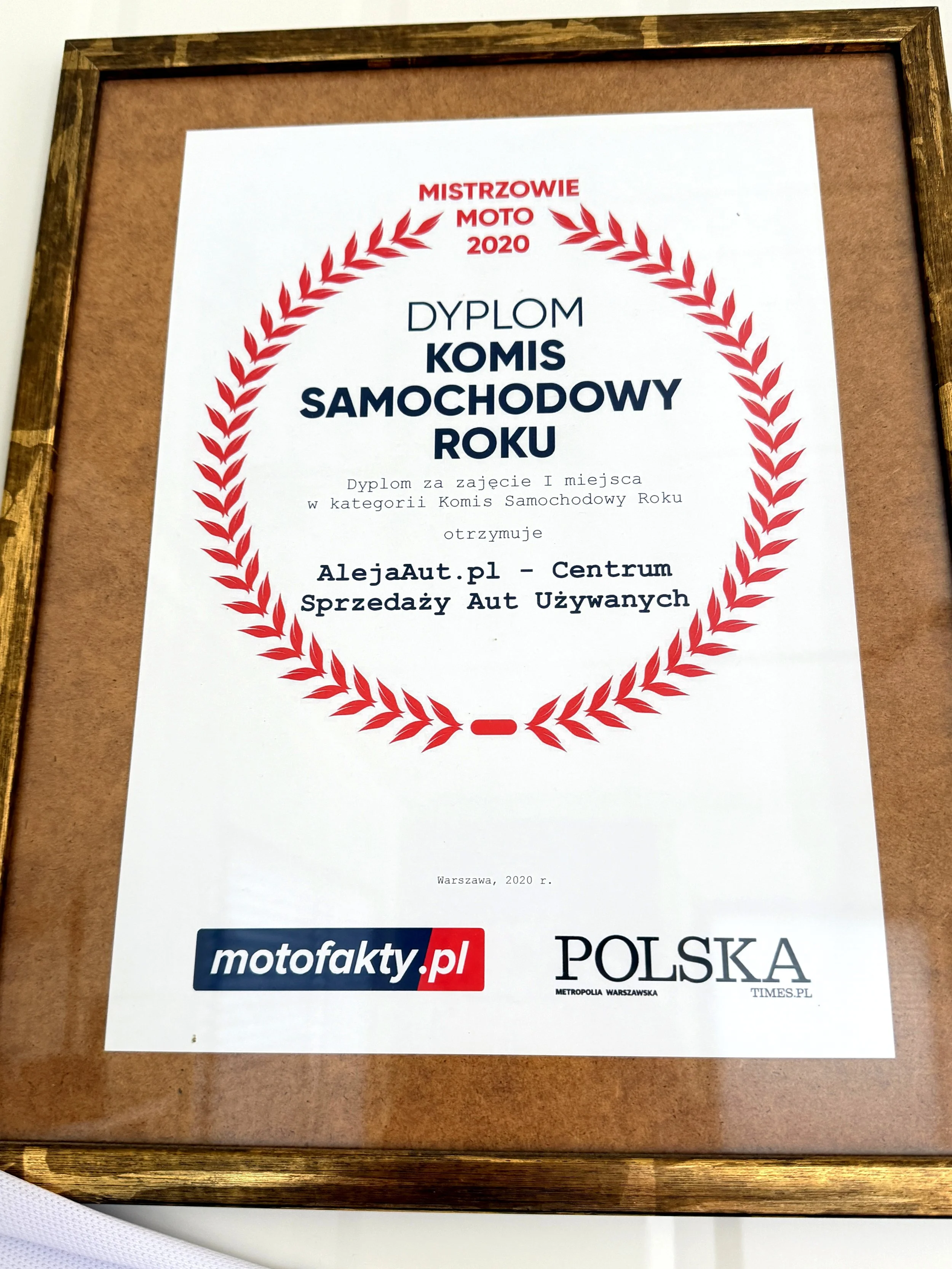 Framed certificate with red laurel wreath design at the top, written in Polish, awarded to a car sales center in Warsaw in 2020 for being the best used car dealership of the year.