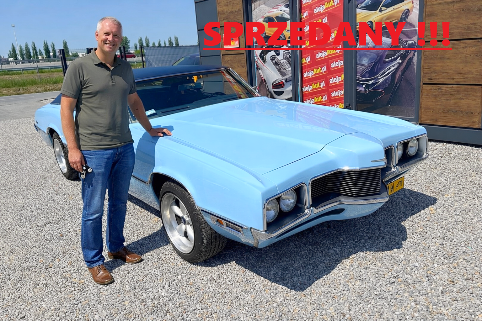A man standing next to a vintage light blue muscle car with a man holding a key, outside a car dealership with a sign that says sold in Polish.