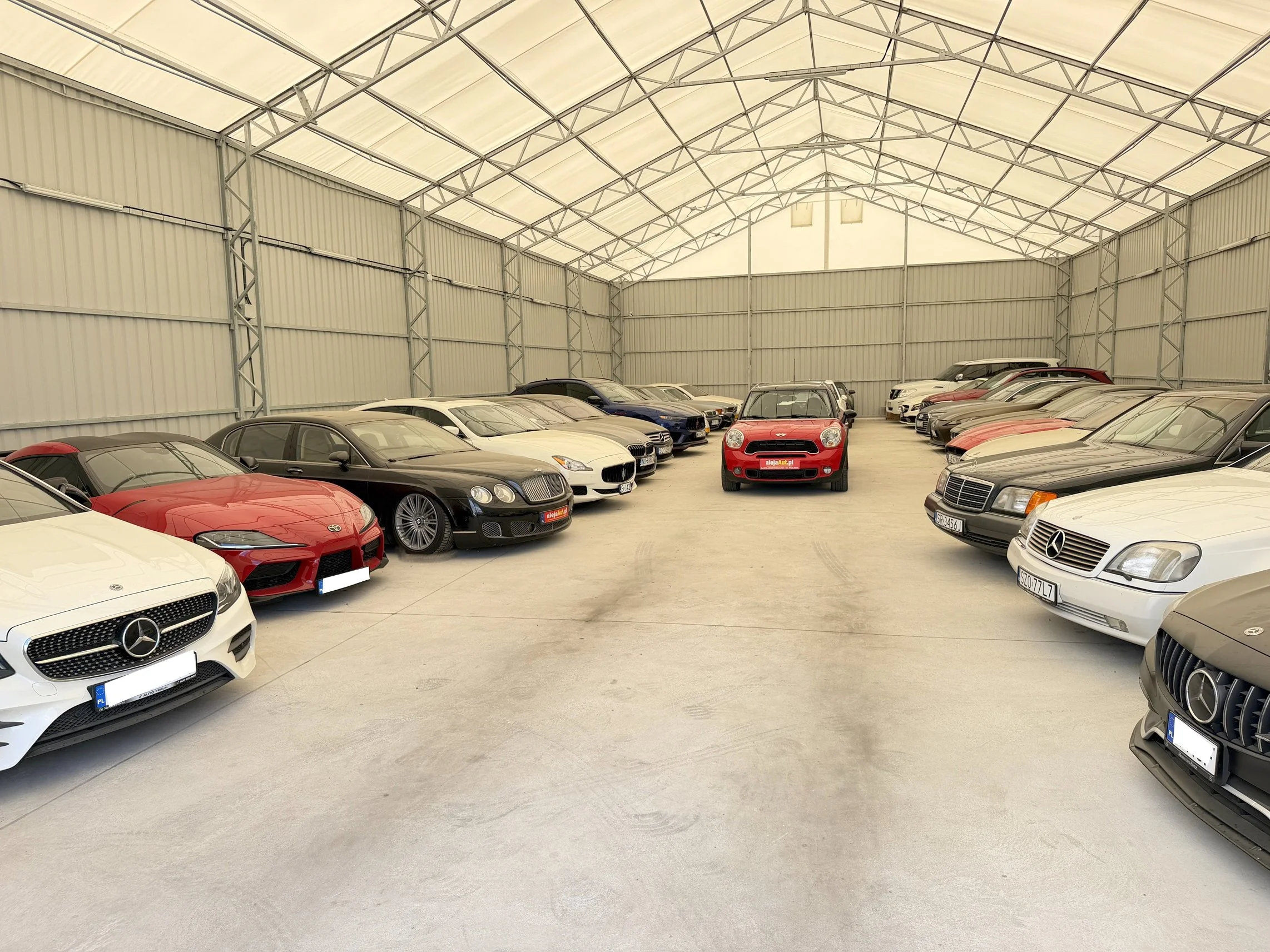 Indoor car storage with numerous luxury vehicles including Mercedes-Benz, Bentley, and Ferrari parked inside a spacious warehouse with a high metal ceiling.