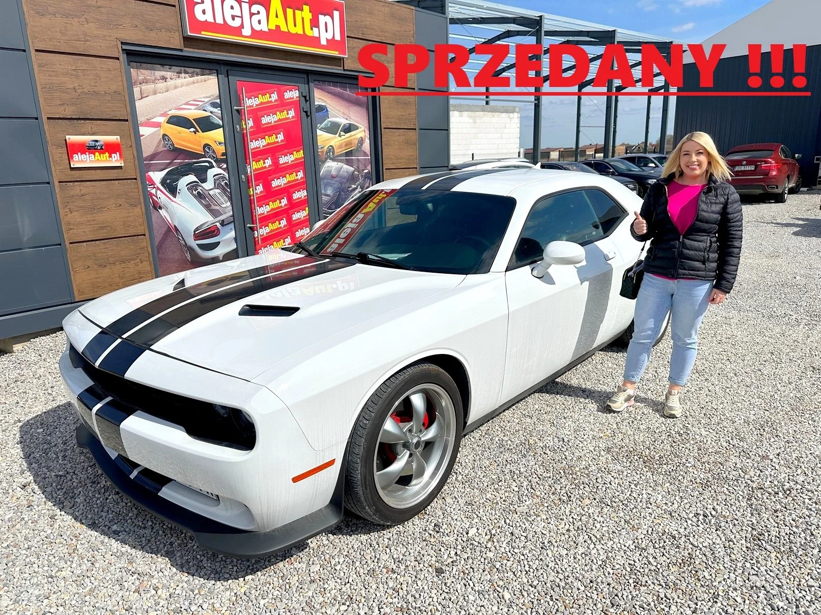A smiling woman in a black jacket and light jeans standing next to a white sports car with black racing stripes outside a dealership with a sign that reads 'alejaAut.pl' and large windows showing images of other cars. The word 'SPRZEDANY!!!' in red capital letters is displayed across the top of the image.