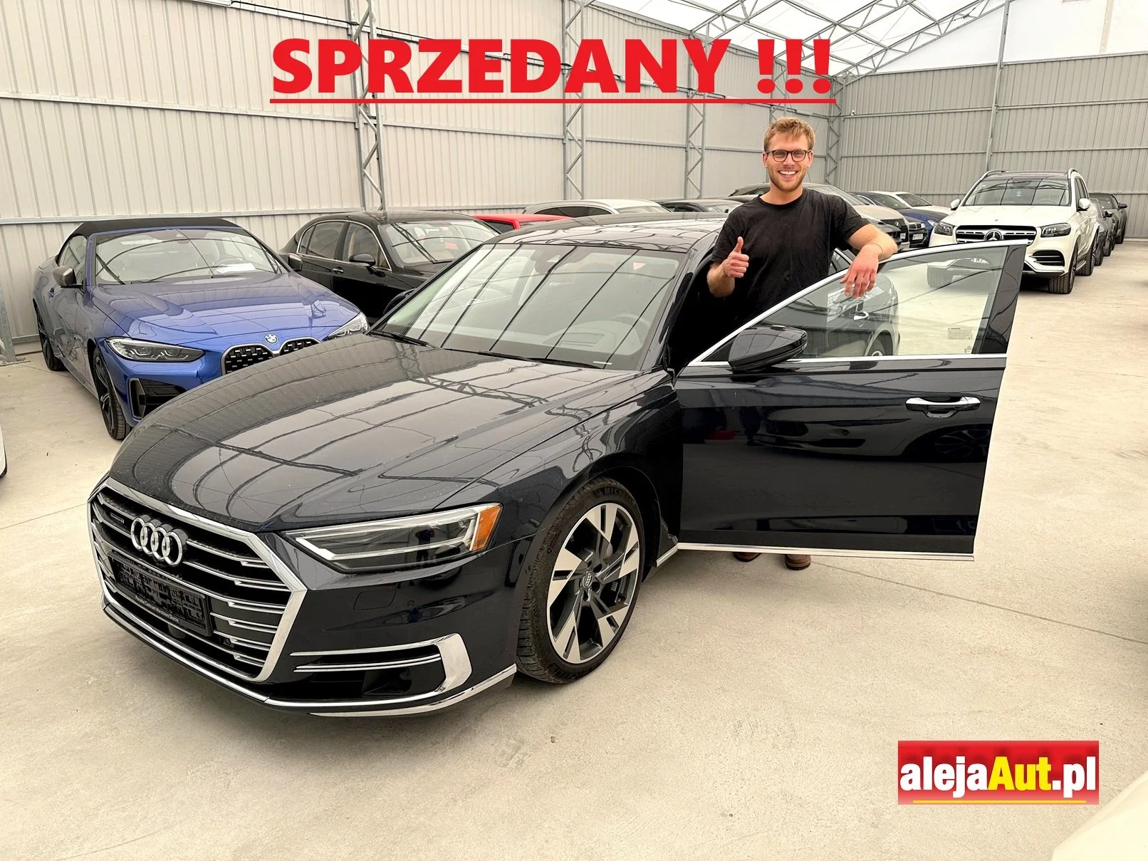 A man standing beside a black Audi car with the door open in a warehouse filled with other cars. The man is smiling and giving a thumbs-up, with the words 'SPRZEDANY!!!' in red text at the top and the logo 'alejaAut.pl' at the bottom right corner.