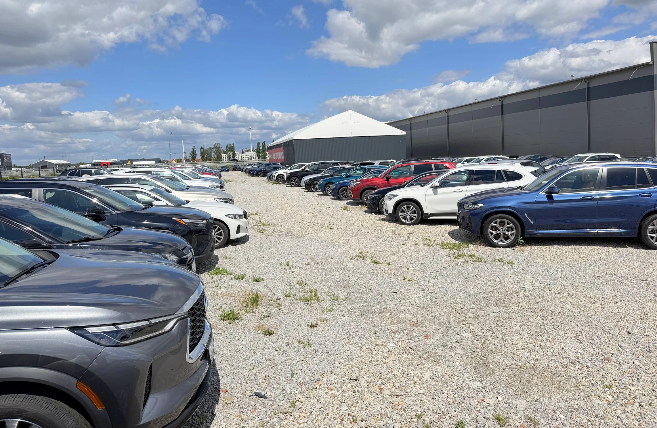 A parking lot filled with various cars in front of large industrial buildings under a partly cloudy sky.