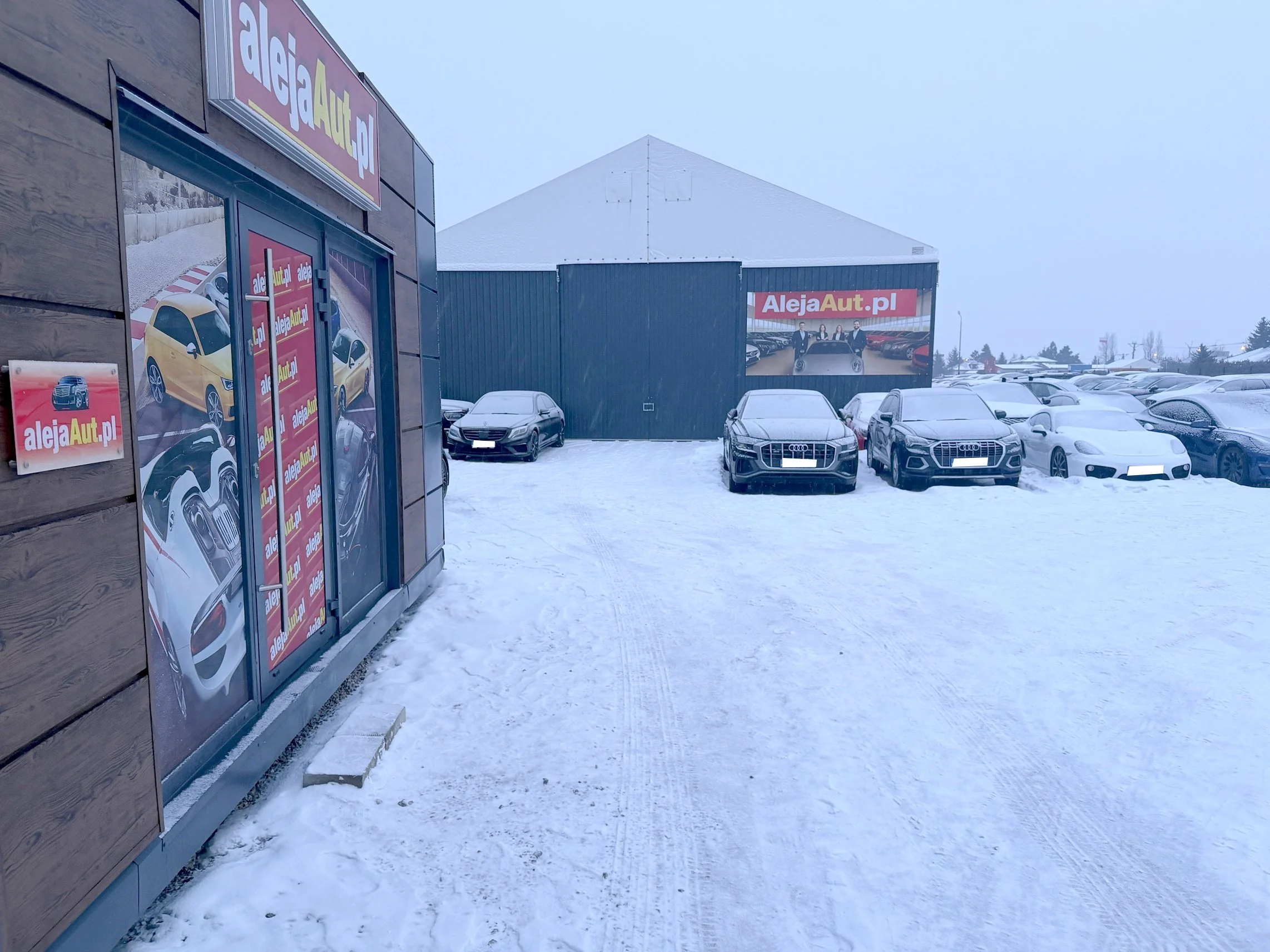 Snow-covered parking lot outside car dealership with cars parked, large signs displaying cars and the website alejaAuto.pl, building with wood and metal siding, overcast sky.