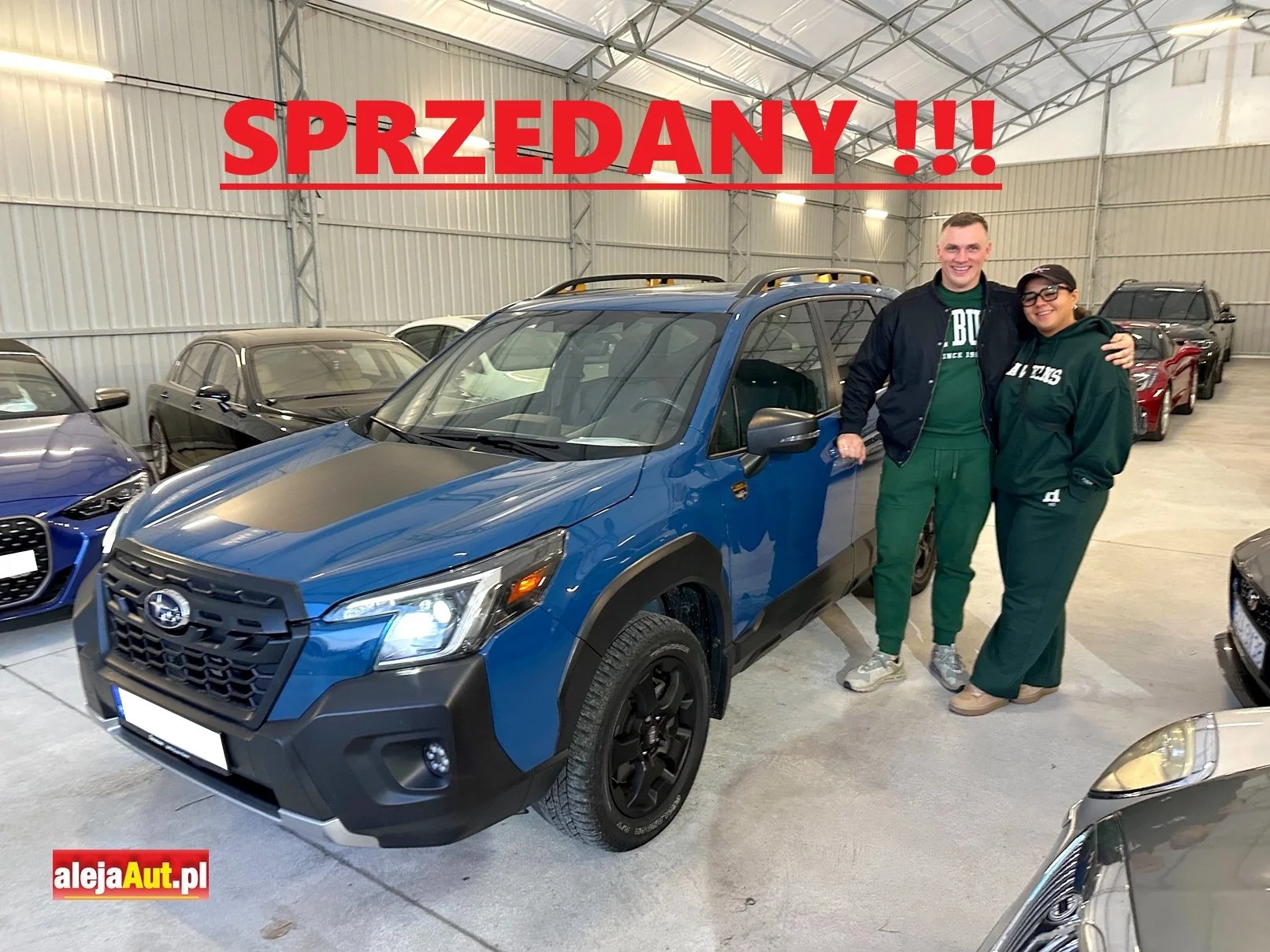 A man and a woman standing together in a garage or showroom with a blue Subaru car behind them, surrounded by other cars. The woman and man are smiling, and the man has his arm around the woman. There is a large red banner with the word 'SPRZEDANY!!!' in Polish across the top and a red and yellow logo with the text 'alejaAut.pl' in the bottom left corner.