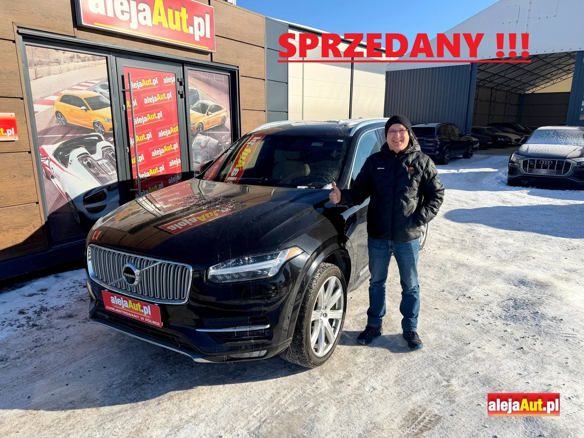 A man standing next to a black Volvo SUV, giving a thumbs-up in front of a car dealership with a sign that says alejaAut.pl and a large 'Sold' sign in red with exclamation marks. The dealership has a window displaying images of various cars, and there are several parked cars on a snowy lot.