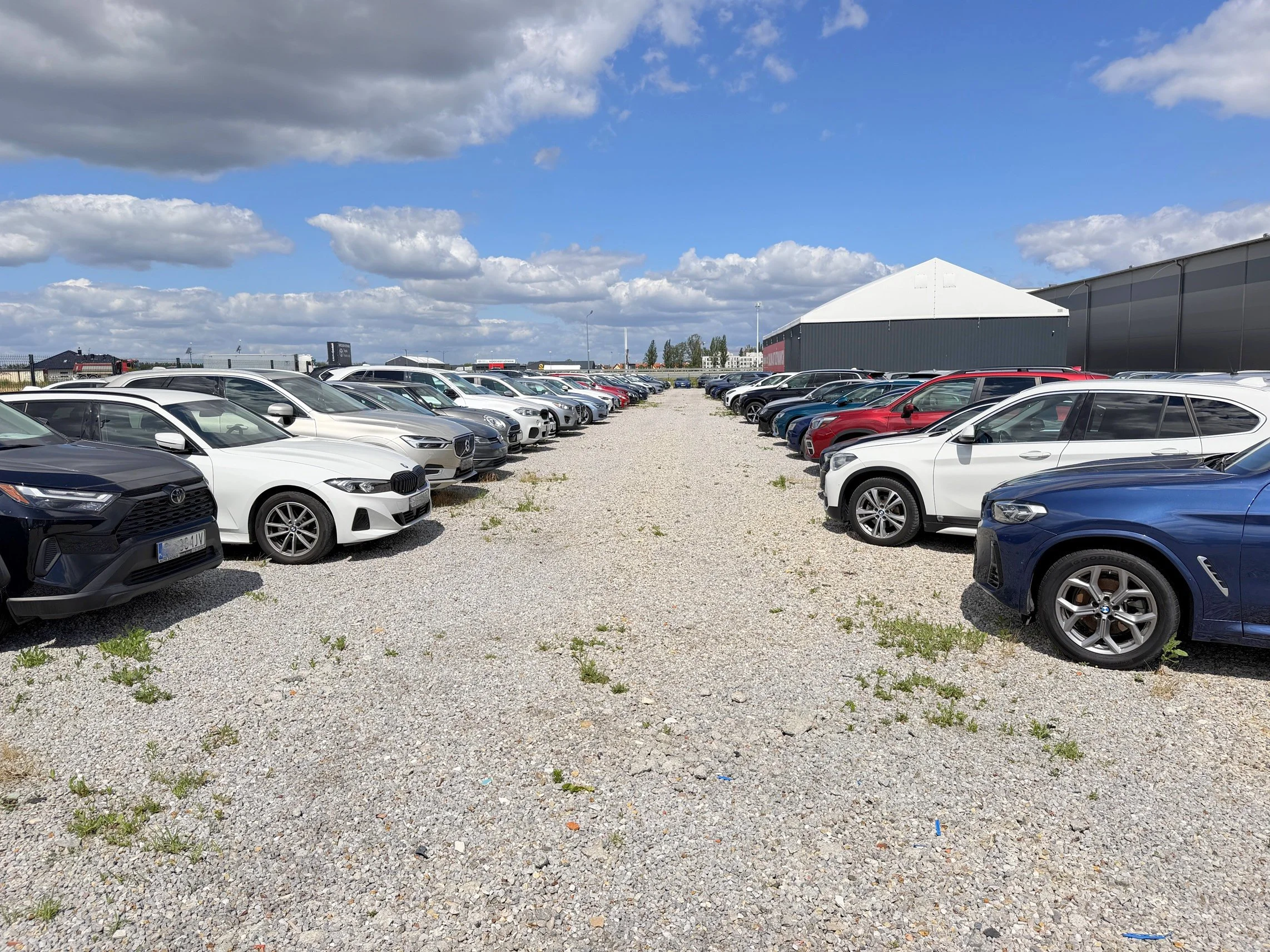 A large outdoor parking lot filled with numerous modern cars, including brands like Mercedes-Benz and Audi, under a partly cloudy sky with white clouds and blue patches, surrounded by industrial buildings and a fence in the background.