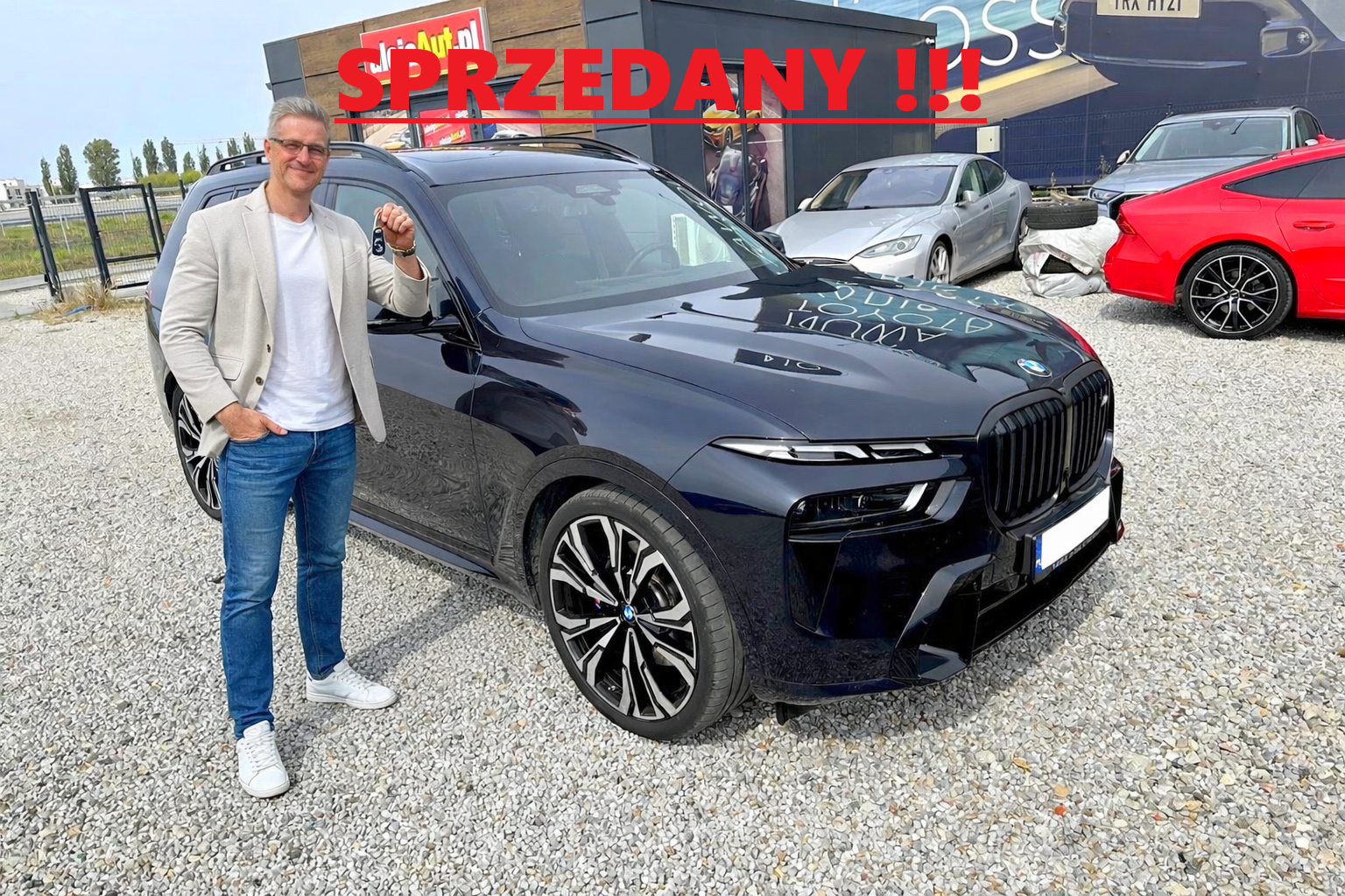 A man in a beige blazer, white t-shirt, blue jeans, and white sneakers standing next to a black BMW SUV with a car key in hand. The car has a 'sold' sign in red with the word 'SPRZEDANY' overlaid in red text. Several other cars are visible in the background, along with a building and a gravel parking area.