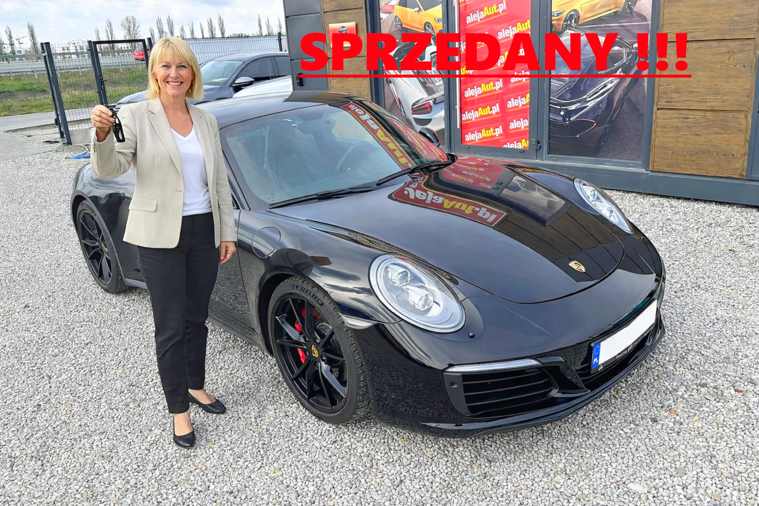 A woman standing next to a black sports car, holding car keys and smiling. The car has the Porsche logo and red brake calipers. There are cars visible in the background and a sign with red text that reads 'SPRZEDANY!!!'.