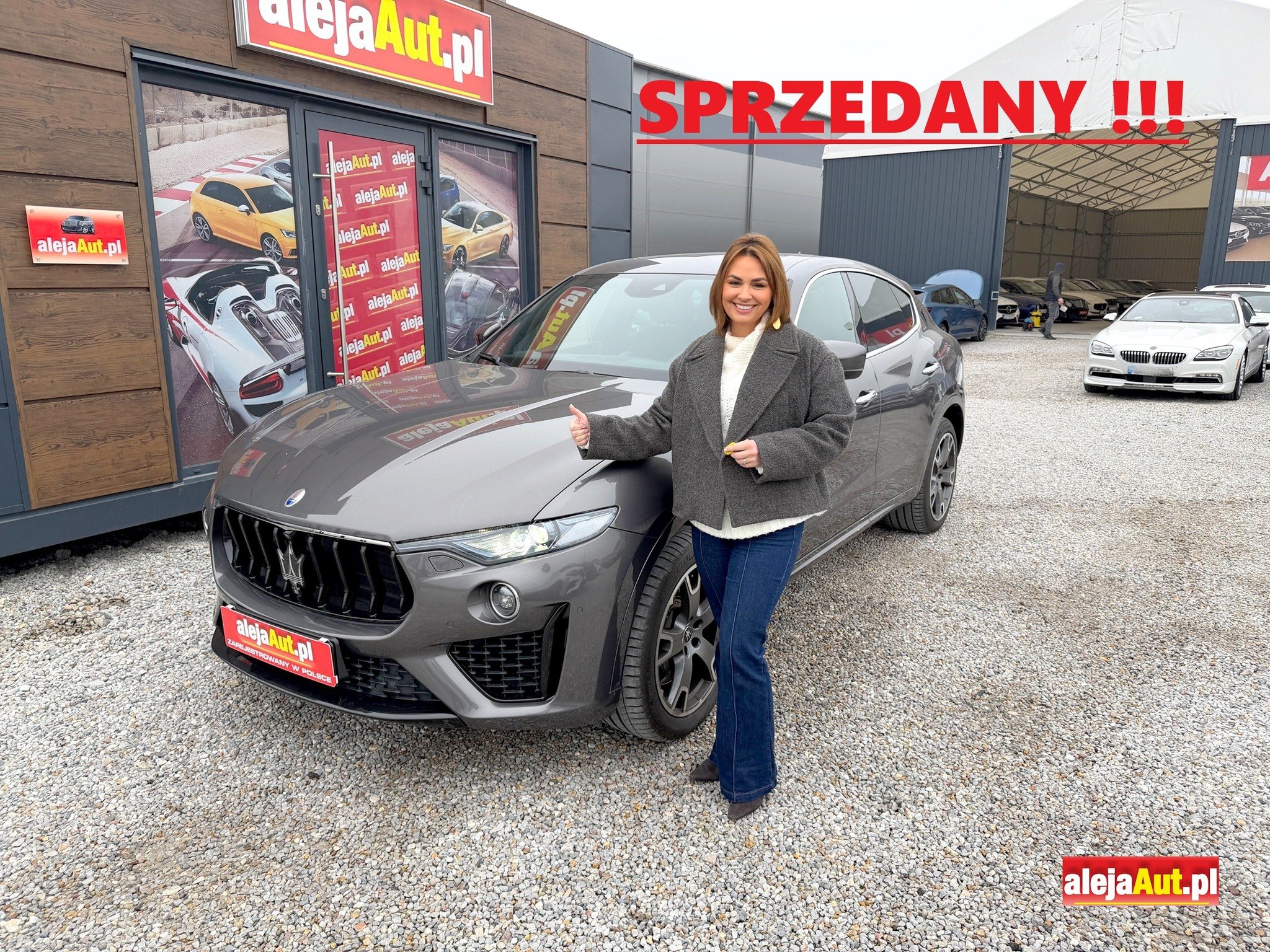 A woman standing next to a gray Maserati car in a car dealership lot, smiling and giving a thumbs up. There are other cars and a building with signage in the background, with a large red banner saying "SPRZEDANY !!!" indicating the car is sold.
