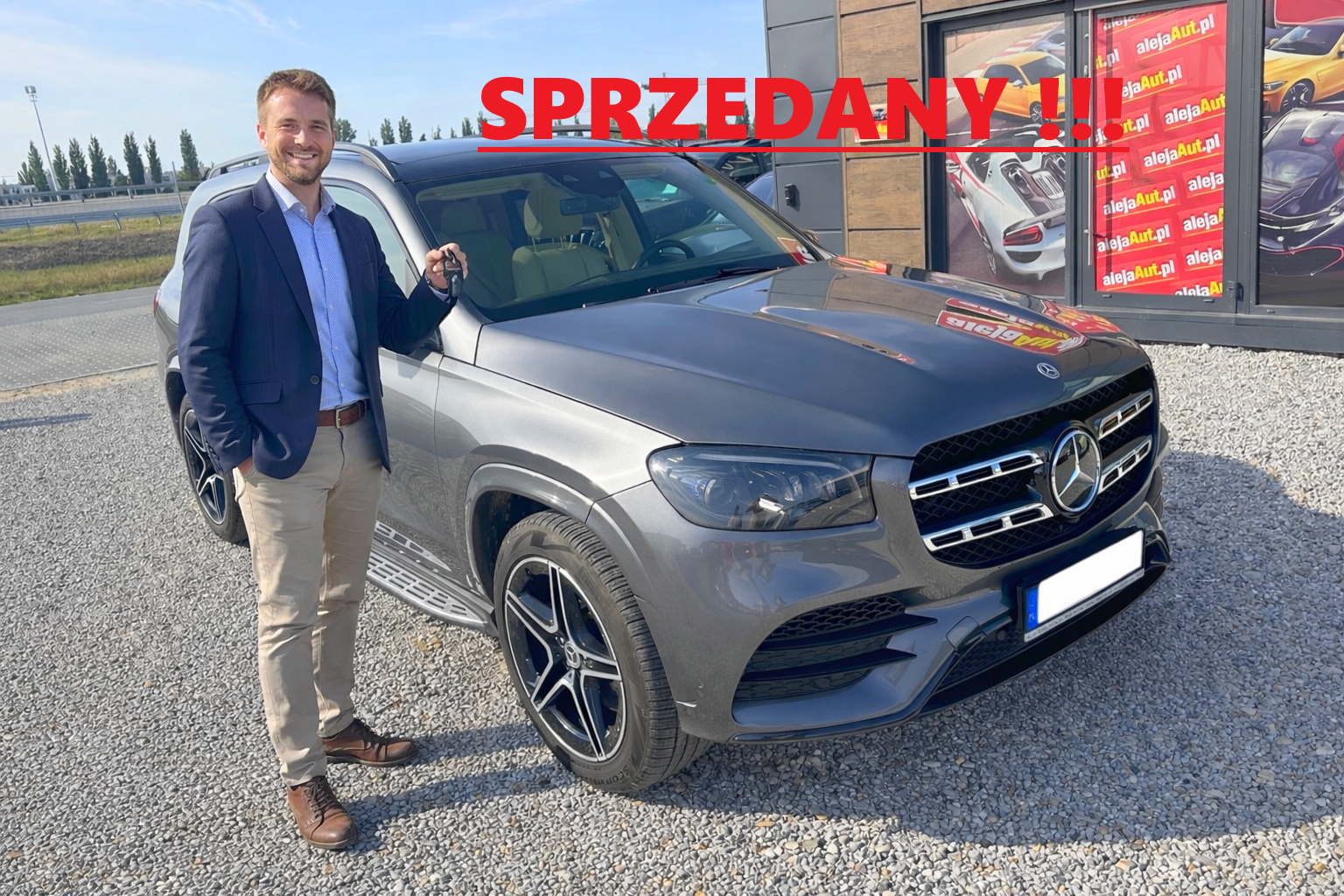 A man in a blue blazer, light blue shirt, beige pants, and brown shoes standing next to a silver Mercedes-Benz SUV, holding car keys, outside a car dealership with a sign reading "SPRZEDANY!!" in red across the top of the image.