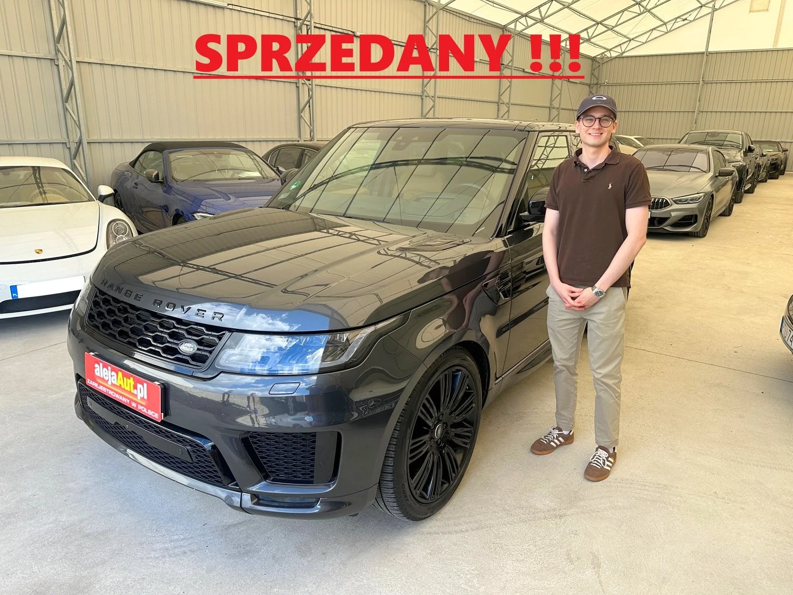 A man standing next to a black Range Rover in a showroom filled with luxury cars, with a red sign reading 'SPRZEDANY!!!' above him.