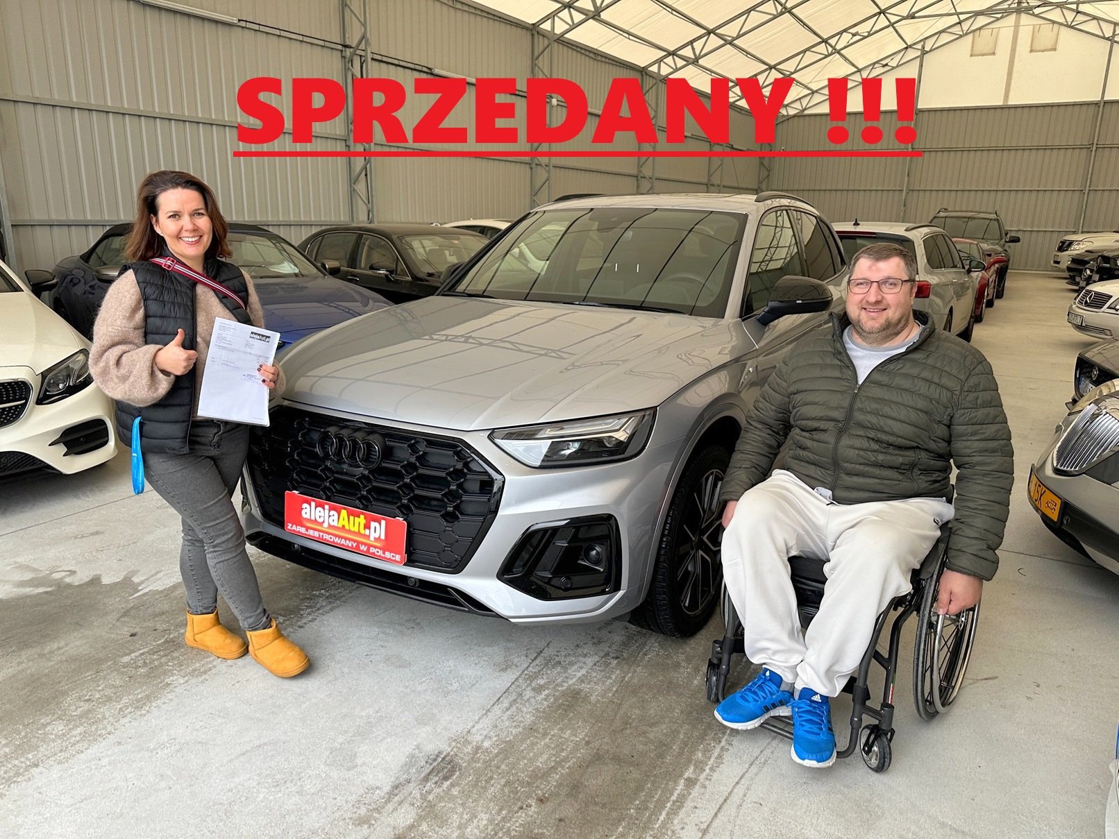 A woman standing next to a silver car holding a document and giving a thumbs-up, and a man in a wheelchair sitting beside the car, both smiling inside a car dealership. The car has a red license plate with the website 'alejaAuto.pl' and the word 'SPZEDANY' with three exclamation marks written in large red letters across the top of the image.