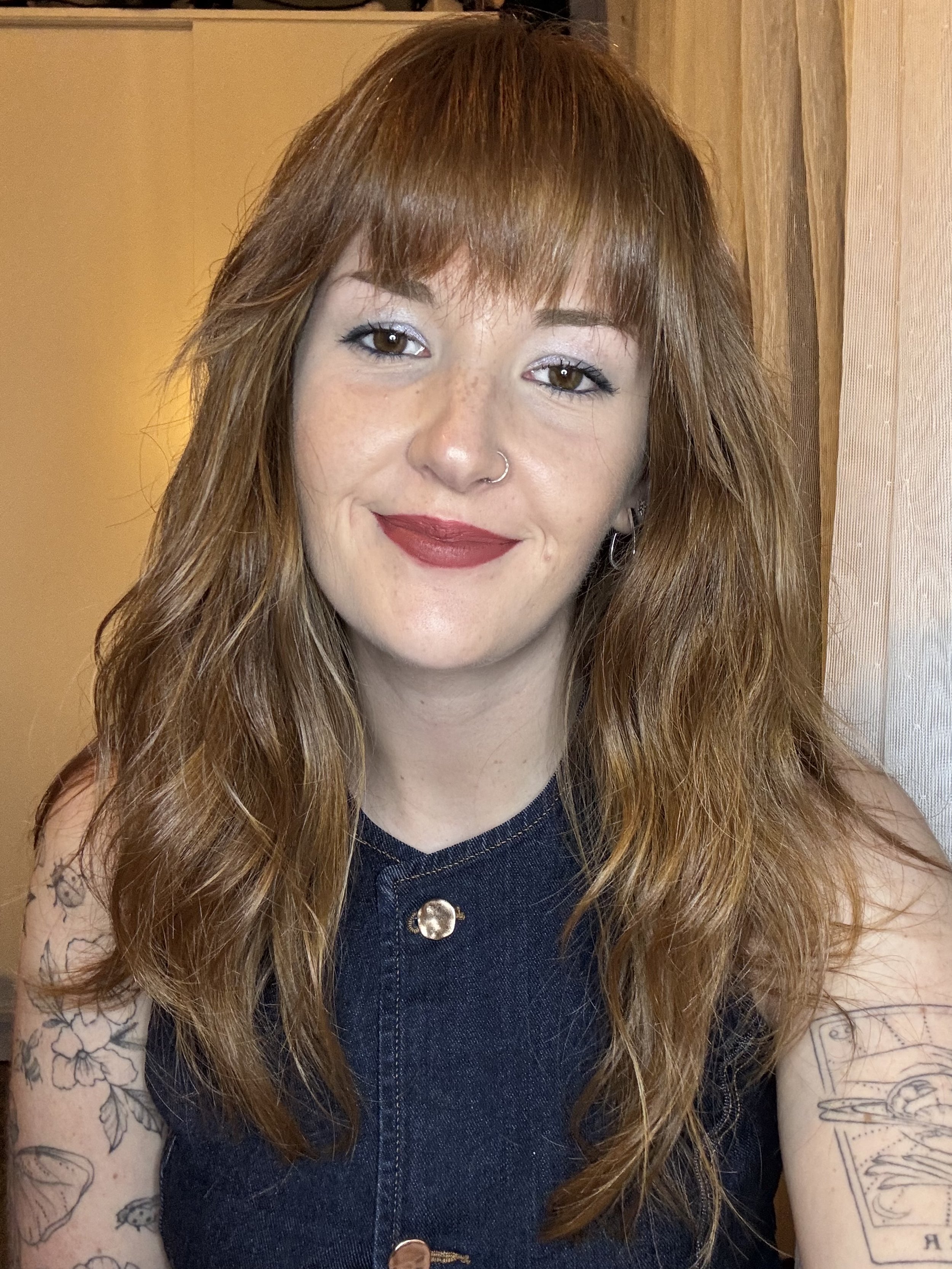 A young woman with long, wavy, red hair and fair skin, smiling, wearing a dark sleeveless denim top, with tattoos on her arms, and a nose ring, inside a room with wooden walls.