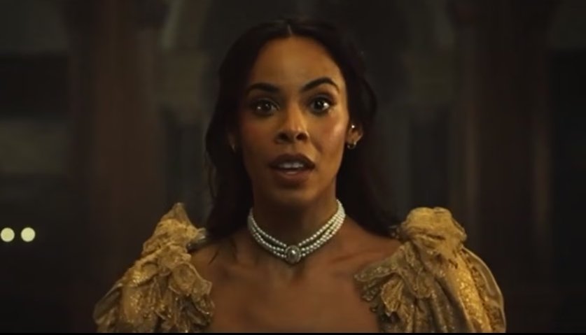 A woman with dark hair wearing a ornate gold dress and a pearl necklace, standing in a dimly lit room.
