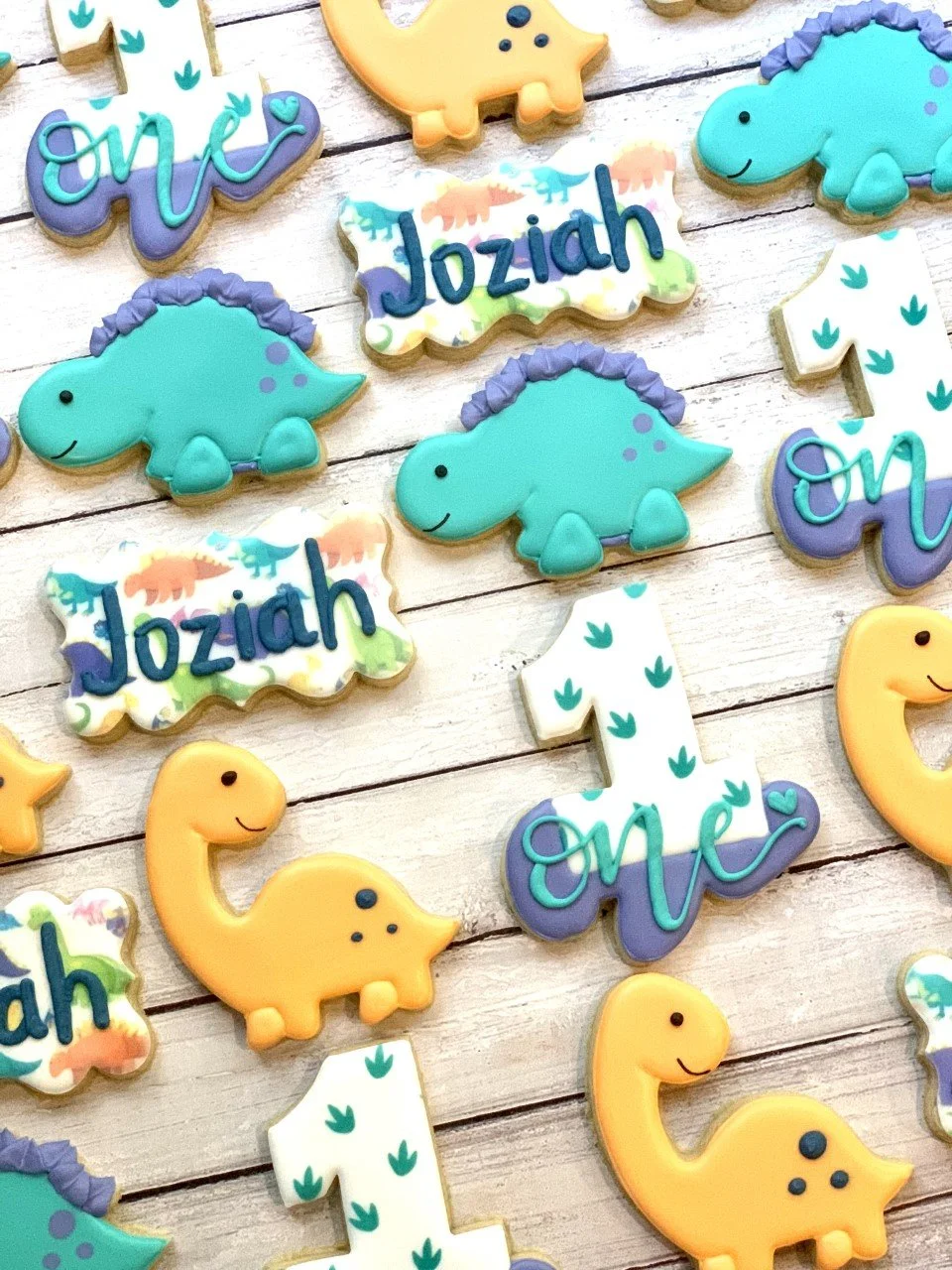 Colorful dinosaur-themed cookies with shapes like dinosaurs, the number one, and personalized name. Decorated with pastel colors and patterns on a wooden surface.