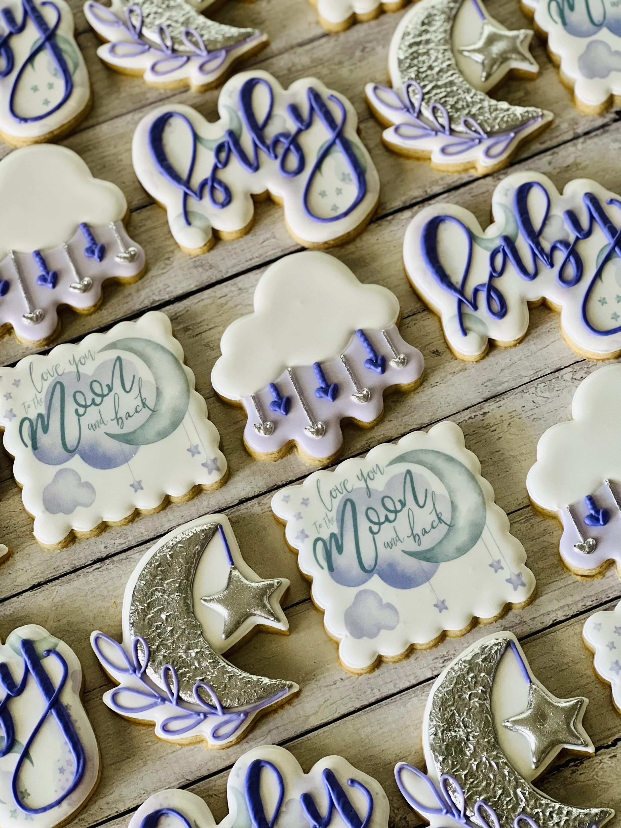 Assorted decorated cookies with pastel and celestial theme, featuring designs of clouds, moons, stars, and words "baby" and "love you to the moon and back," in silver and lavender colors, on a wooden surface.