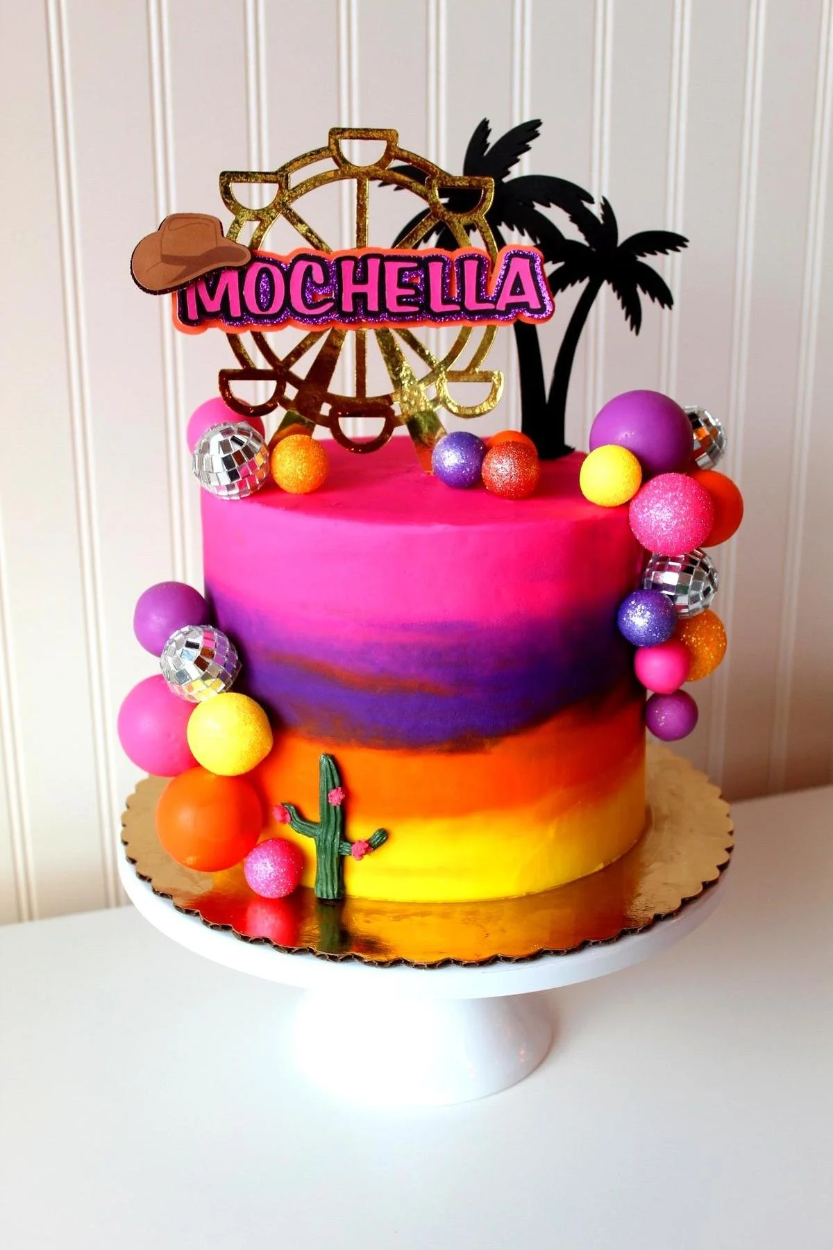 Colorful cake with "MOCHELLA" sign, Ferris wheel and palm tree toppers, decorated with vibrant balls and cactus.