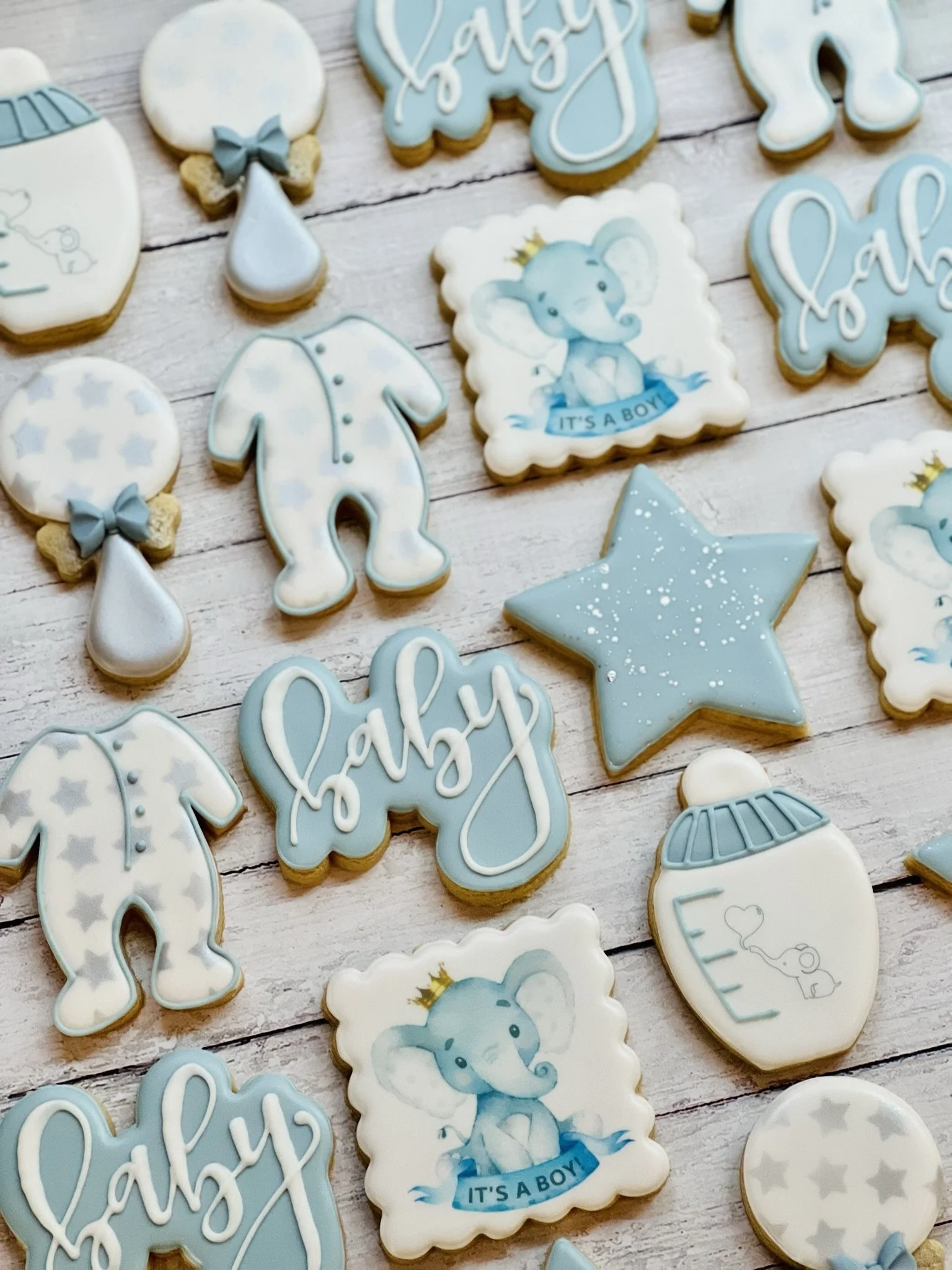 Assorted baby-themed cookies in blue and white, including shapes of onesies, baby bottles, balloons, stars, cursive 'baby' text, and a square cookie with an elephant illustration labeled 'It's a boy.'