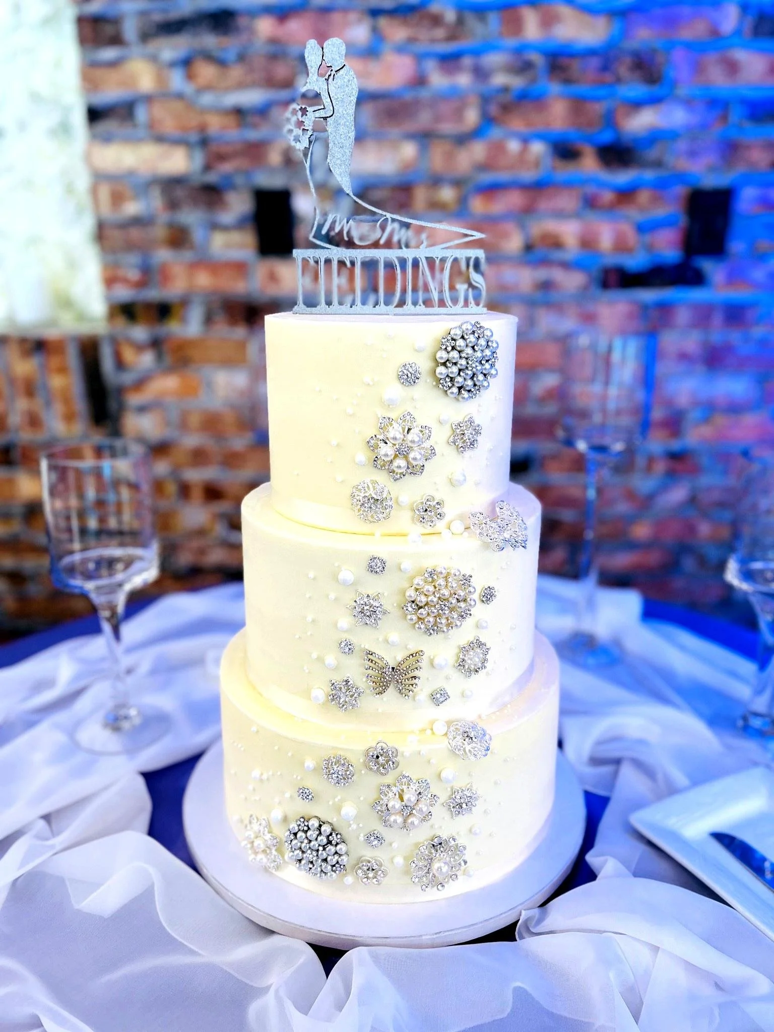 Three-tier white wedding cake with pearl and jewel decorations, topped with a silhouette cake topper on a table with white fabric.