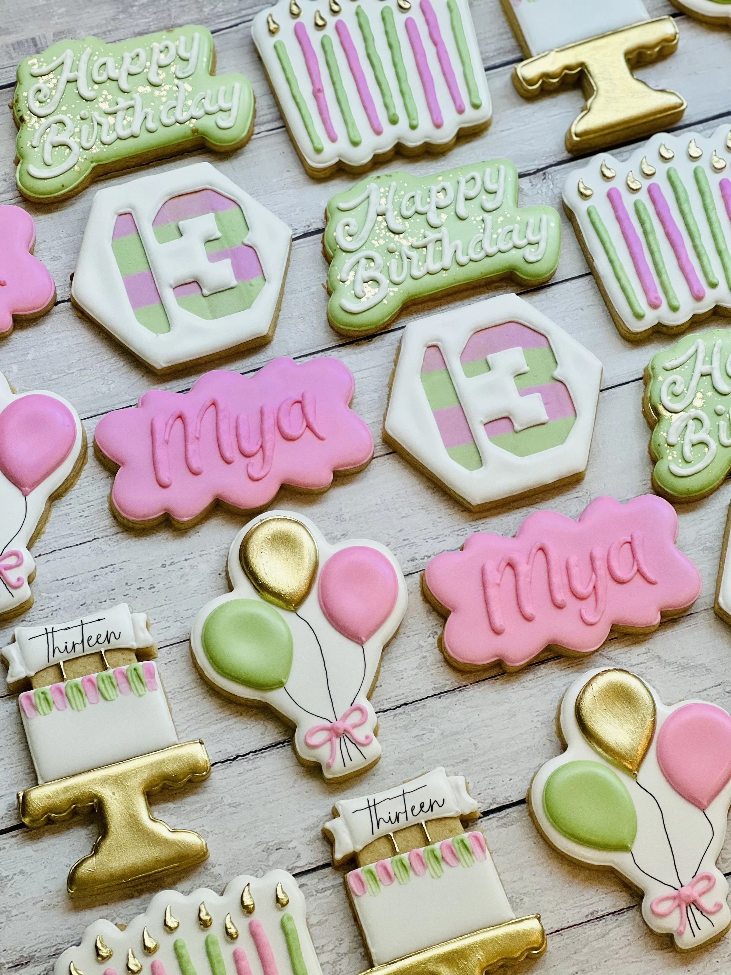 Decorative birthday cookies in pastel colors shaped like cakes, balloons, and signs with 'Happy Birthday,' '13,' and 'Mya' written on them. The cookies feature pink, green, and gold accents.