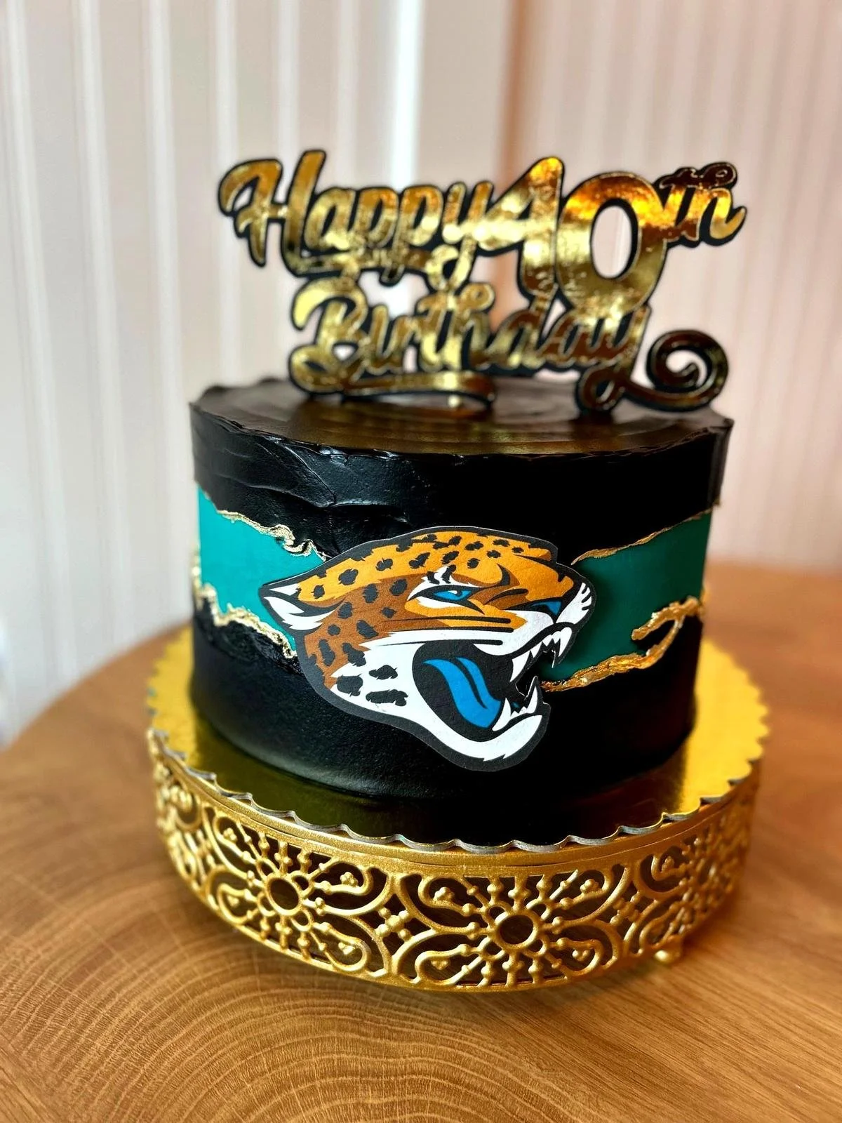 A black and teal cake with a gold topper that reads "Happy 40th Birthday," featuring a jaguar logo. The cake is placed on an ornate gold stand.