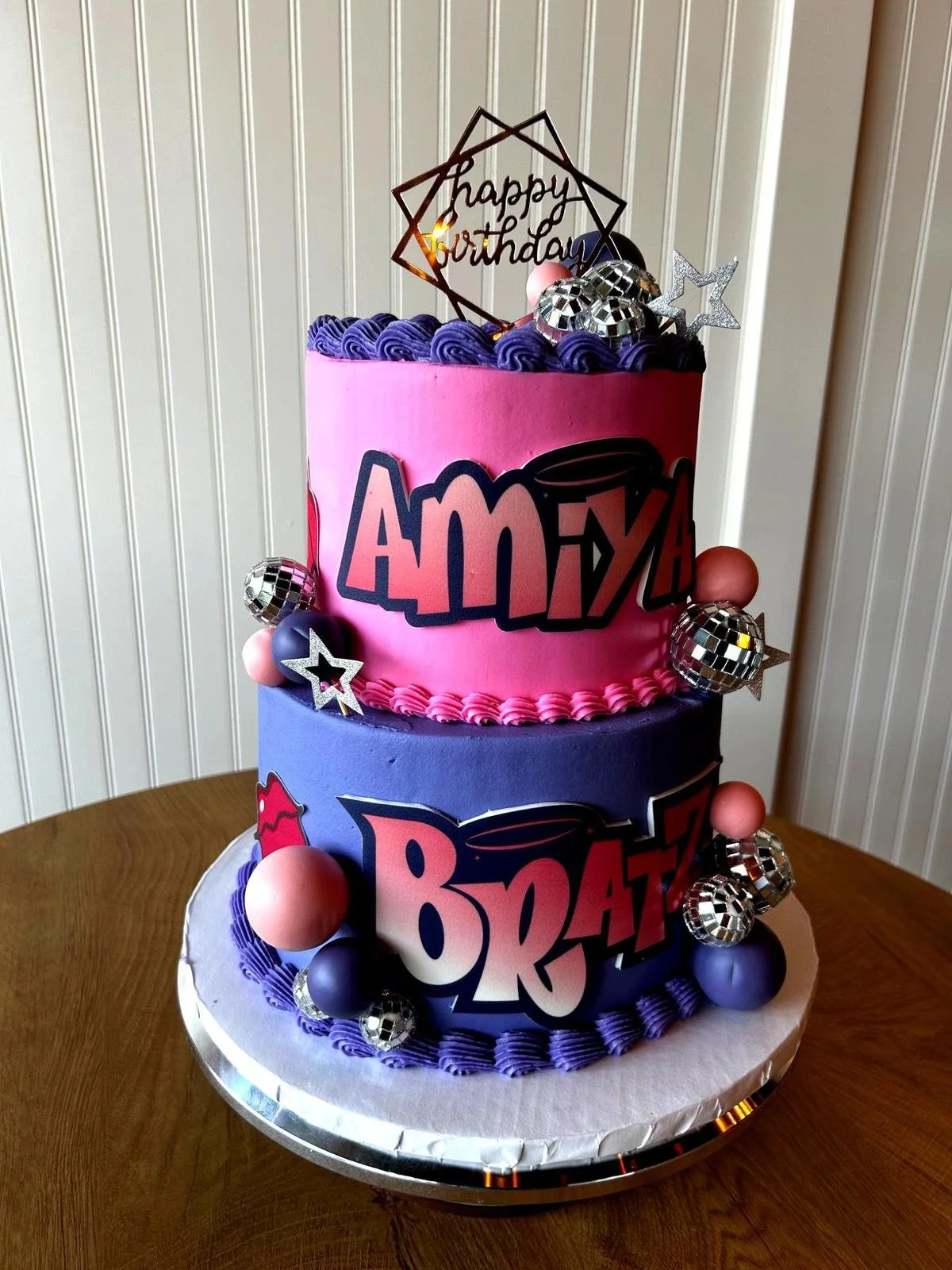 Two-tier birthday cake with pink and purple icing, featuring "Amiyah" and "Bratz" logos, decorated with disco balls and stars, topped with "happy birthday" sign.