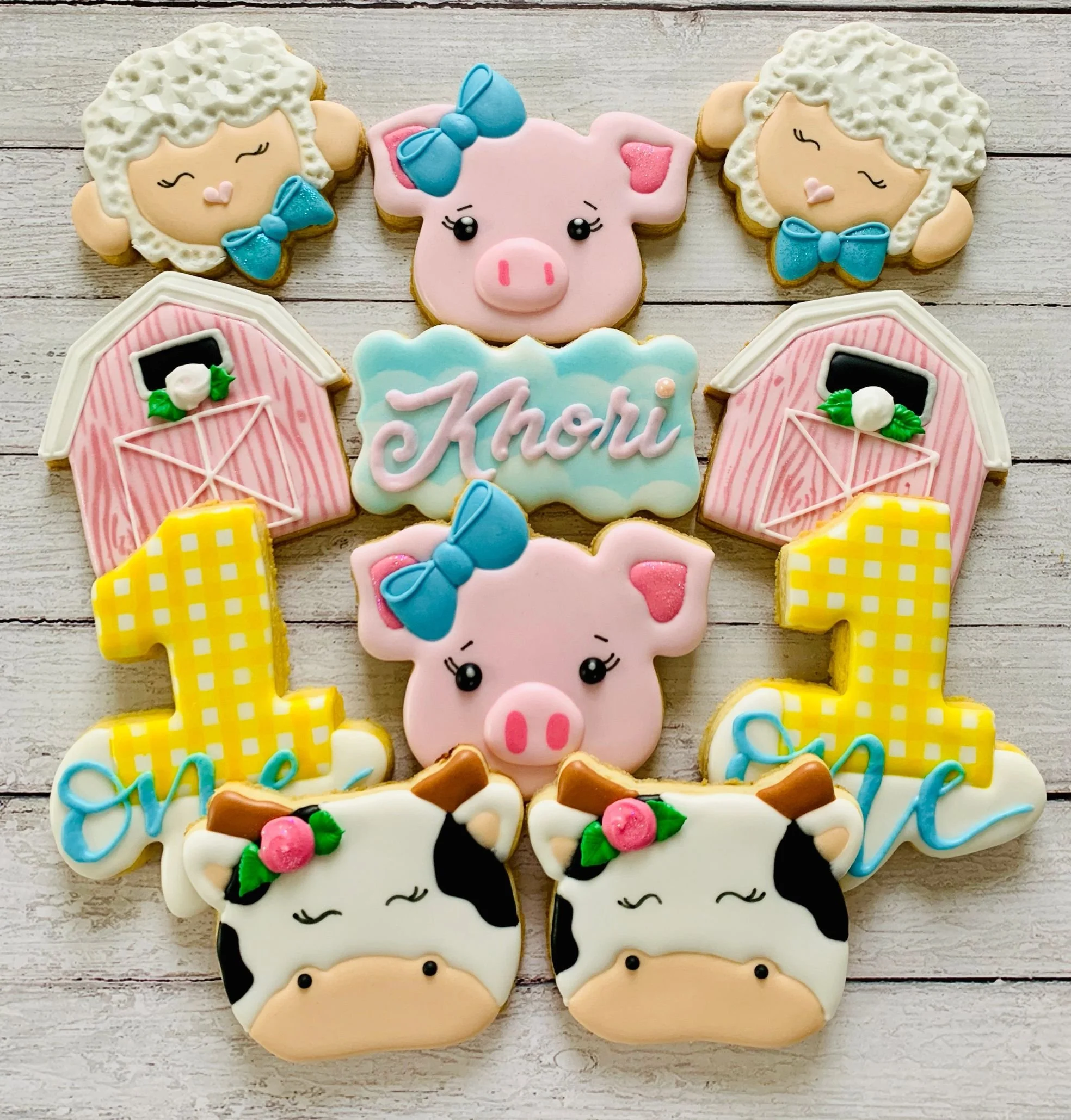 Farm-themed cookies with animal designs, including sheep, pigs, cows, barns, and number ones, displayed on a wooden background.