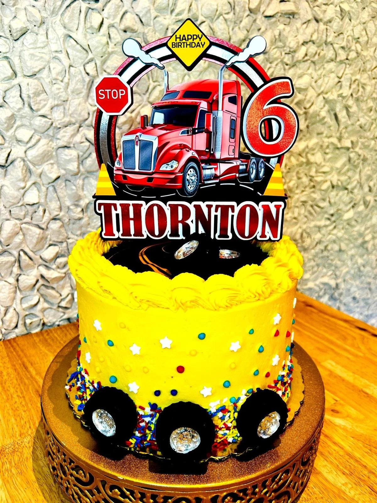 Colorful birthday cake with a truck theme; features a red truck topper, yellow frosting, sprinkles, and a number 6, celebrating a sixth birthday.
