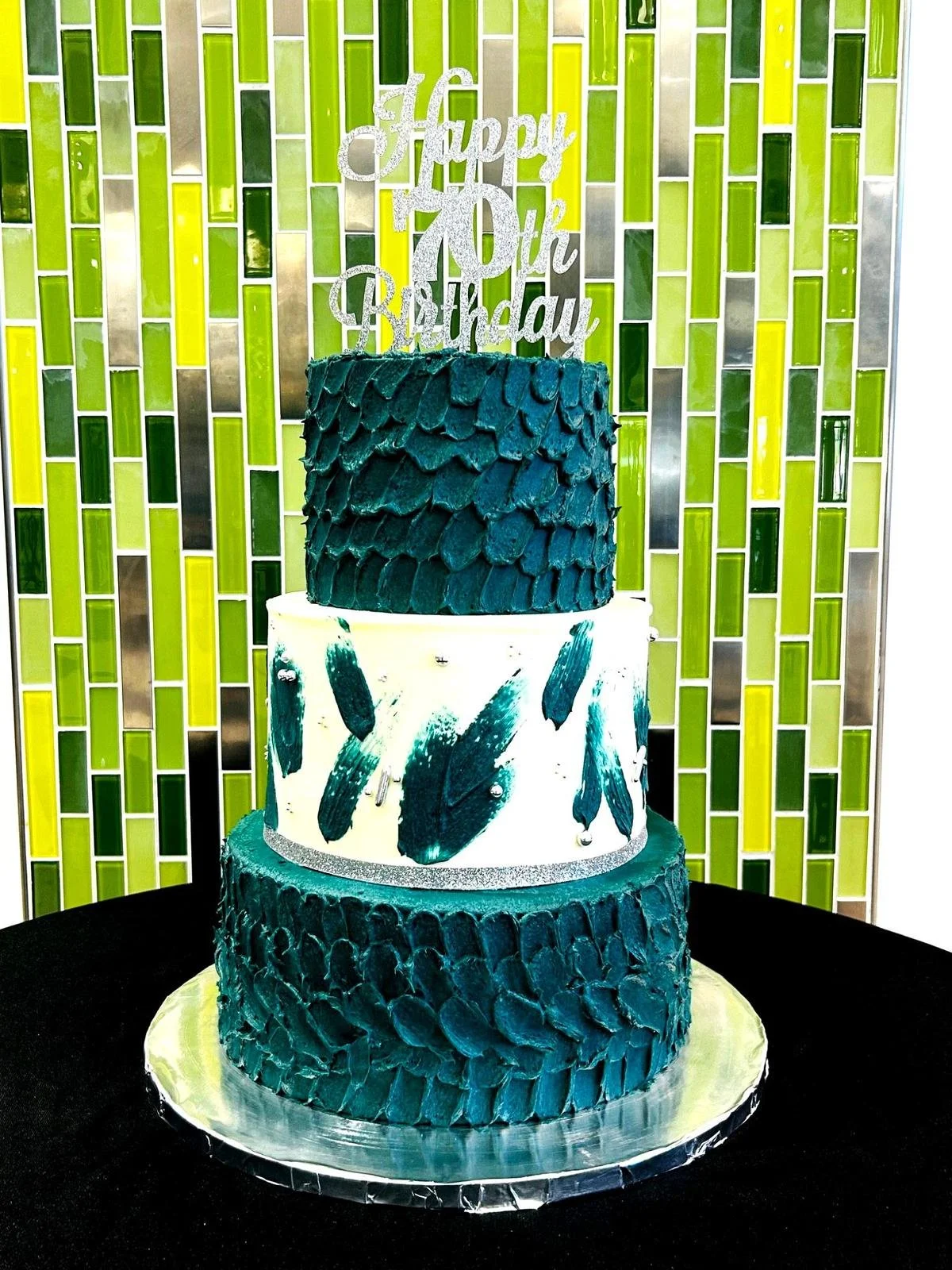 Three-tiered green and white birthday cake with "Happy 70th Birthday" topper, against a green tiled background.