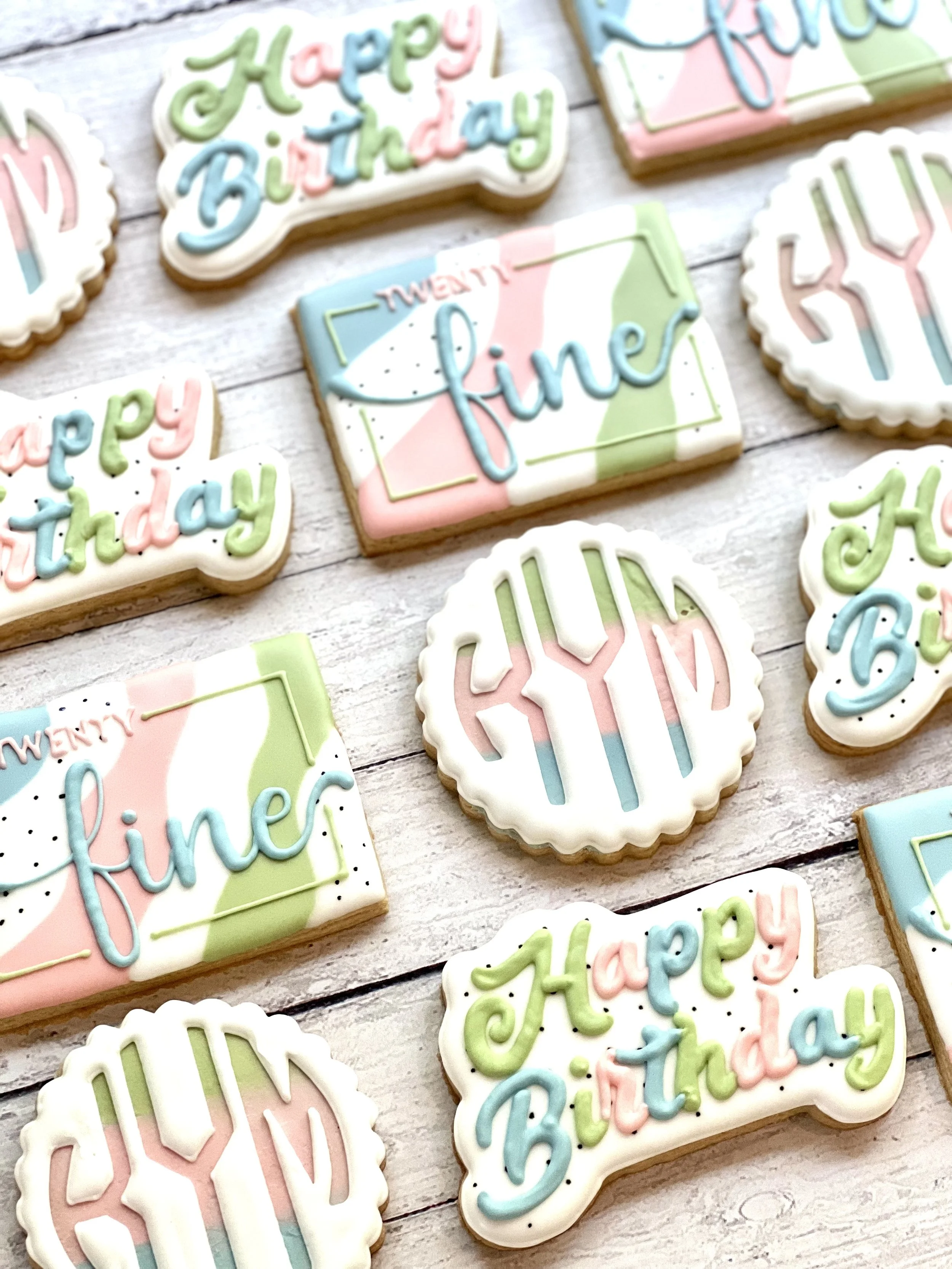 Decorative cookies with pastel icing featuring phrases like "Happy Birthday" and "twenty fine," and initials "KYM" on a wooden surface.