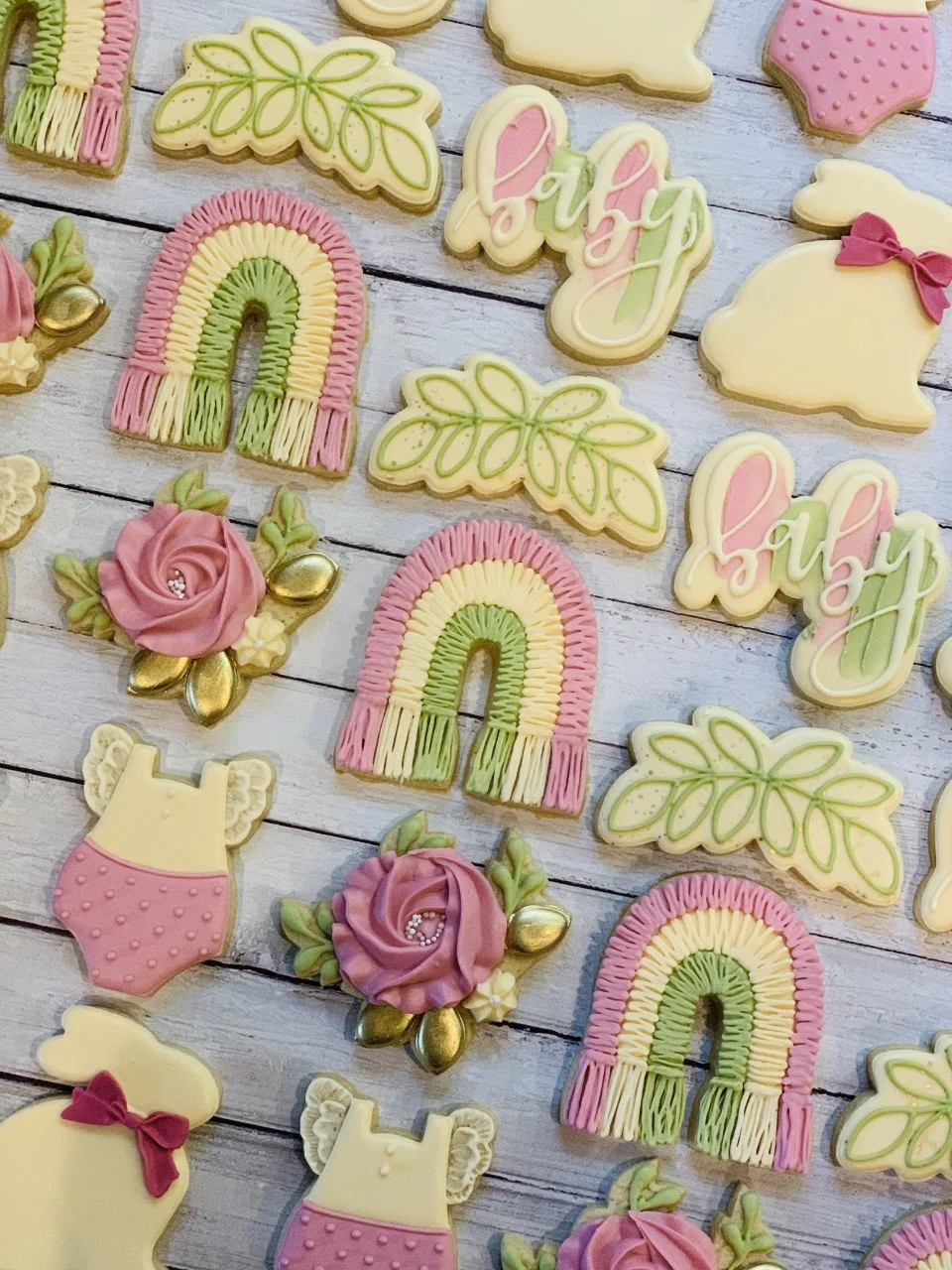 Decorative baby shower cookies with pastel designs including rainbows, roses, baby outfits, leaves, and the word 'baby' on a wooden background.