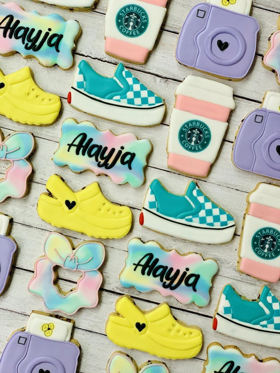 Decorated cookies in various shapes, including cameras, Starbucks cups, sneakers, and sandals, with pastel colors and the name "Alayja" on some.