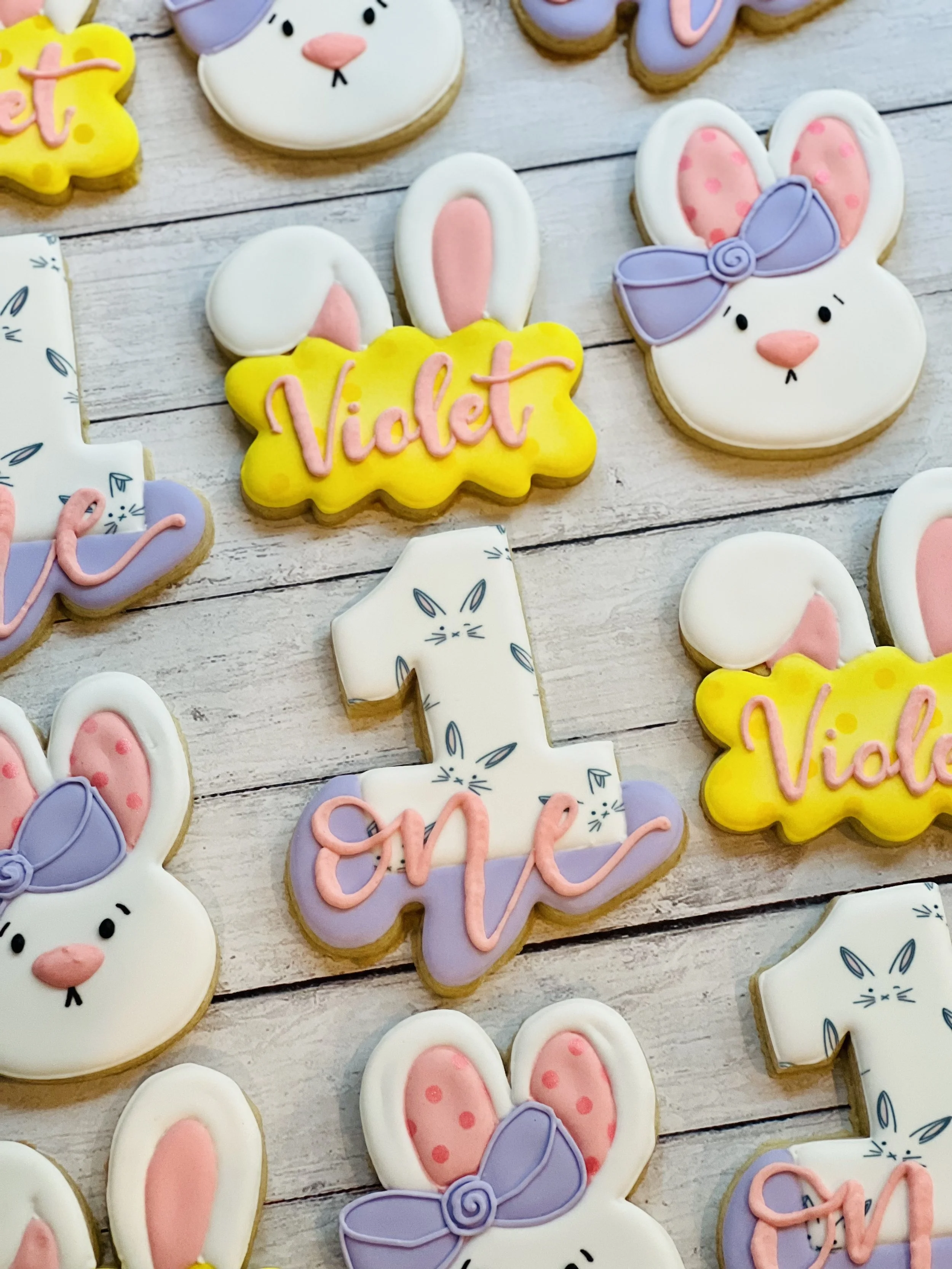 Decorated cookies with rabbit faces and text, including the name "Violet" and the number one, on a wooden surface.
