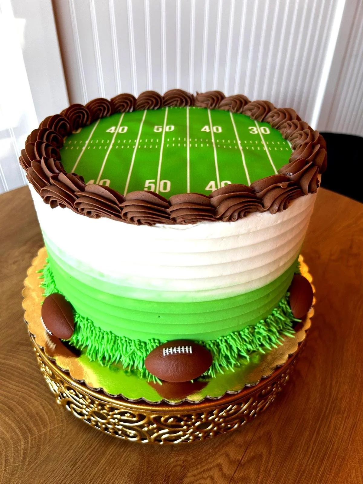 Layered cake with a football field design on top and chocolate football decorations, sitting on a decorative base.