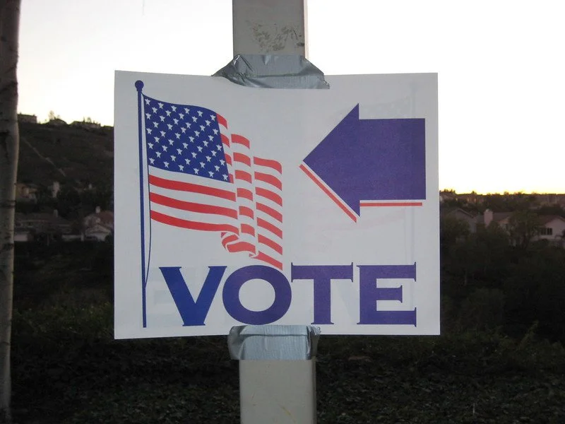 Photo of a sign on a pole with an American flag, arrow pointing left and word "VOTE"