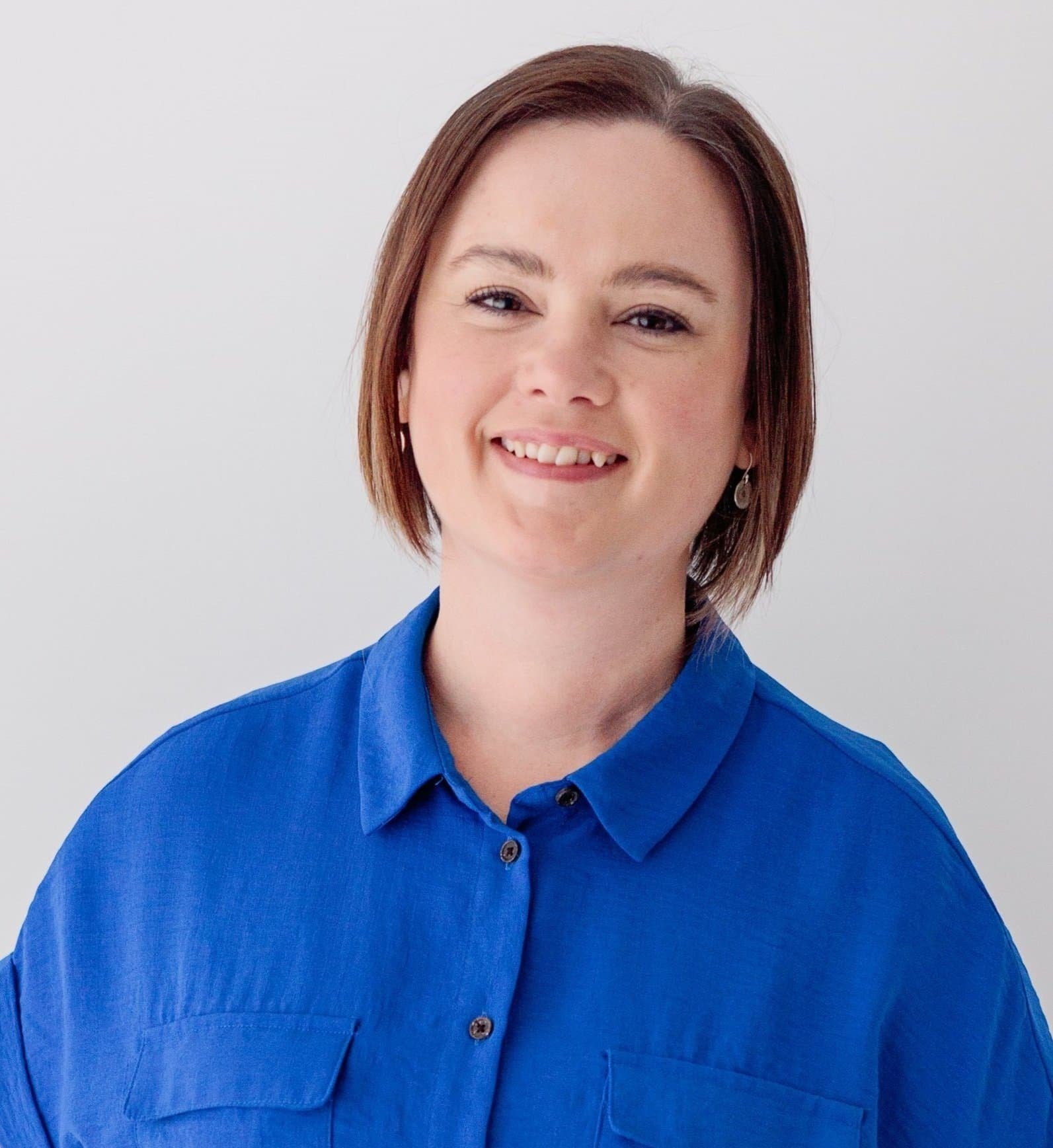 A picture of Louise Hiles. Smiling woman with short brown hair wearing a bright blue button-up shirt, standing in front of a plain white background.